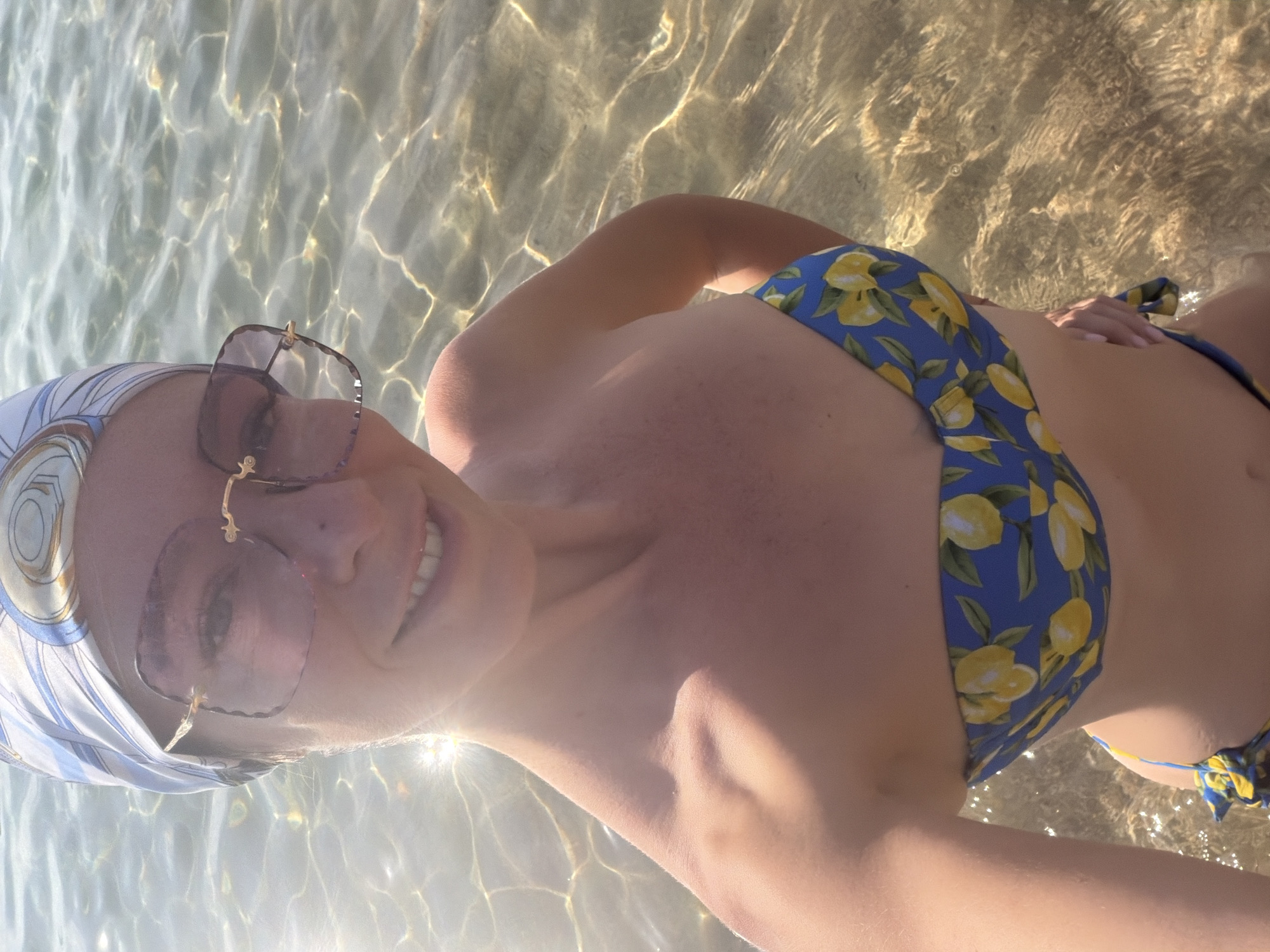A woman wearing a blue bikini with a lemon print and a patterned headscarf stands in shallow, clear water, with sunlight glinting off the surface. She is wearing large, square-shaped sunglasses with rose-tinted lenses. The woman is looking at the camera and smiling. The background is the sea, with the seabed visible beneath the water. The image was taken in Nha Trang, Vietnam.