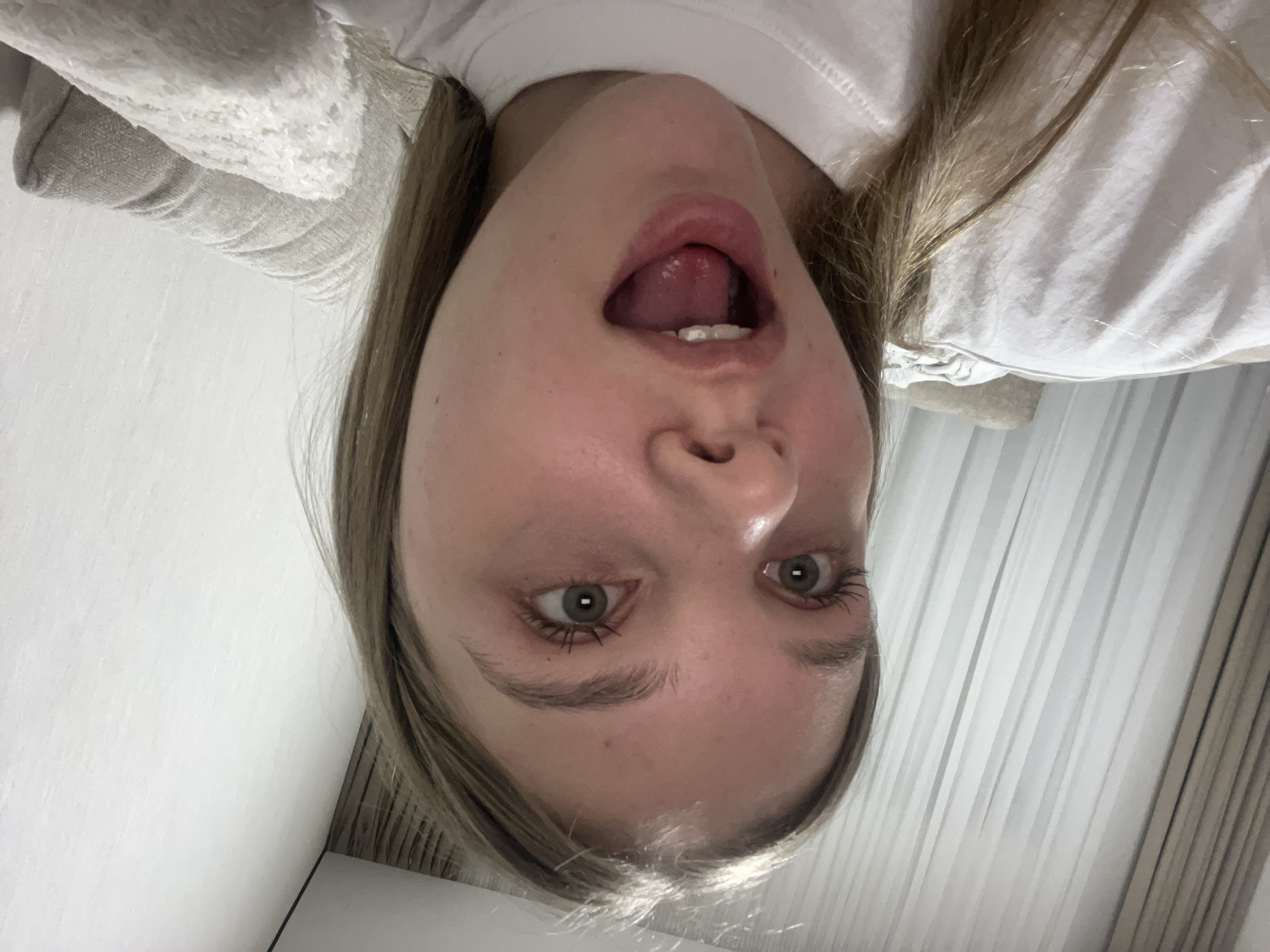 An inverted, close-up selfie captures a young person, likely female, in an indoor setting. The subject has light brown or blonde hair and is wearing a plain white t-shirt or top.
The person's face is central, with wide-open eyes showing light-colored irises and dark pupils. Their eyelashes appear to have mascara. The mouth is open, with the tongue slightly extended and a few front teeth visible, suggesting a playful, surprised, or silly expression. The perspective indicates the subject is lying down and looking up at the camera.
In the background, a plain white wall or ceiling is visible on the left. On the right side, light-colored curtains or blinds with vertical stripes are discernible. A soft, textured fabric, possibly a blanket or part of a sofa, is visible behind the upper left shoulder area. The lighting is ambient and uniform, typical of indoor conditions. There is no visible text in the image. The scene conveys an informal and lighthearted moment, taking place in Votkinsk, Russia.
