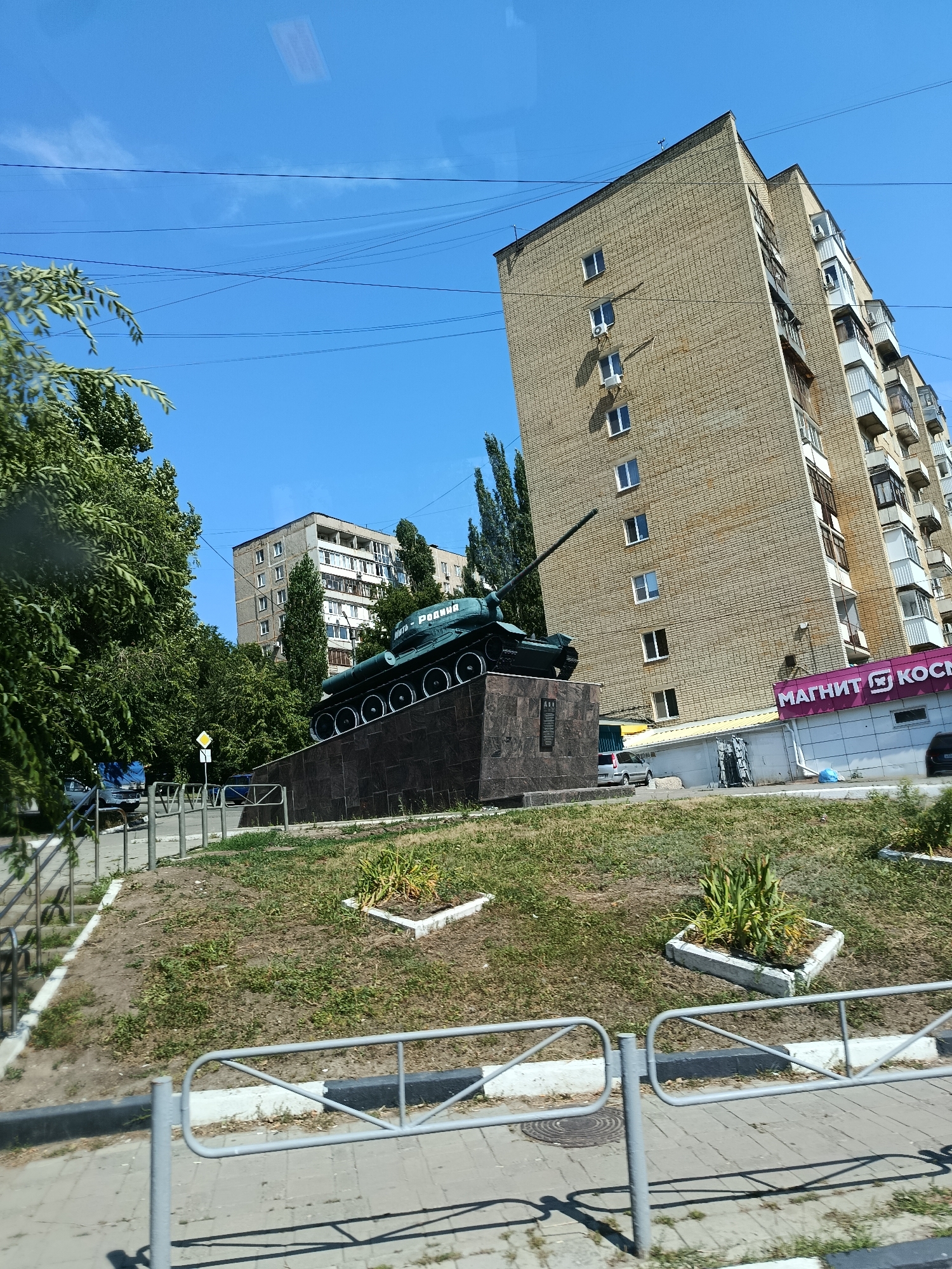 The image shows a monument to a Soviet T-34 tank on a raised platform in the city of Saratov, Russia. The tank is painted dark green and has the inscription "ВЕЛИКАЯ ОТЕЧЕСТВЕННАЯ - РОДИНА" (Great Patriotic War - Motherland) on its side. The monument is located in front of a multi-story brick apartment building. There are trees and a blue sky with some power lines in the background. In the foreground, there is a sidewalk with a metal fence and some greenery. The overall scene depicts an outdoor urban environment.