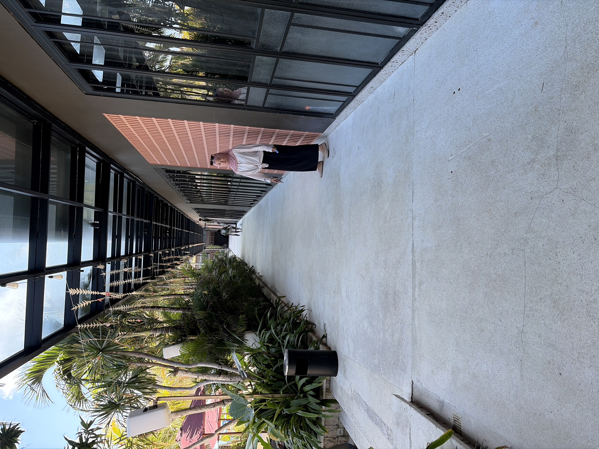A woman wearing a pink hijab, a white long-sleeved blouse, and a black maxi skirt stands on a concrete walkway in Kuta Selatan, Indonesia. The walkway is lined with large windows on the right and lush green foliage on the left. A black trash can is visible near the plants. Overhead, a glass-covered walkway with exposed lighting fixtures creates a structured canopy. In the distance, another person walks away from the camera. The scene appears to be an outdoor commercial or hospitality area.