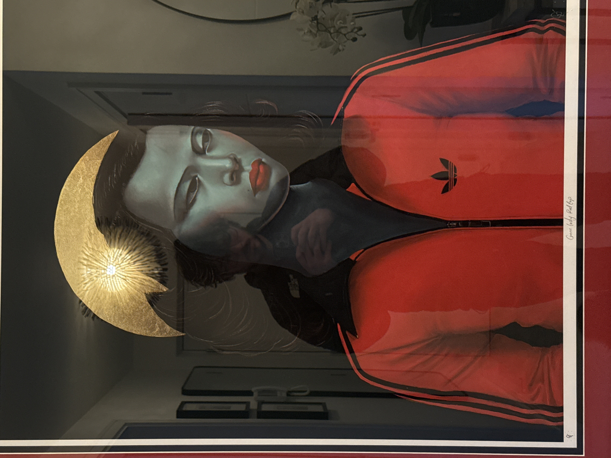 A framed print or painting depicts a woman in an Adidas track jacket with a golden halo. The woman has fair skin, dark hair, and red lipstick. The track jacket is red with black stripes. The golden halo is behind her head and has a radiant design. The print is framed with a white border and a wider red border. The surrounding environment is reflected on the print glass. The location is Unknown, Unknown.