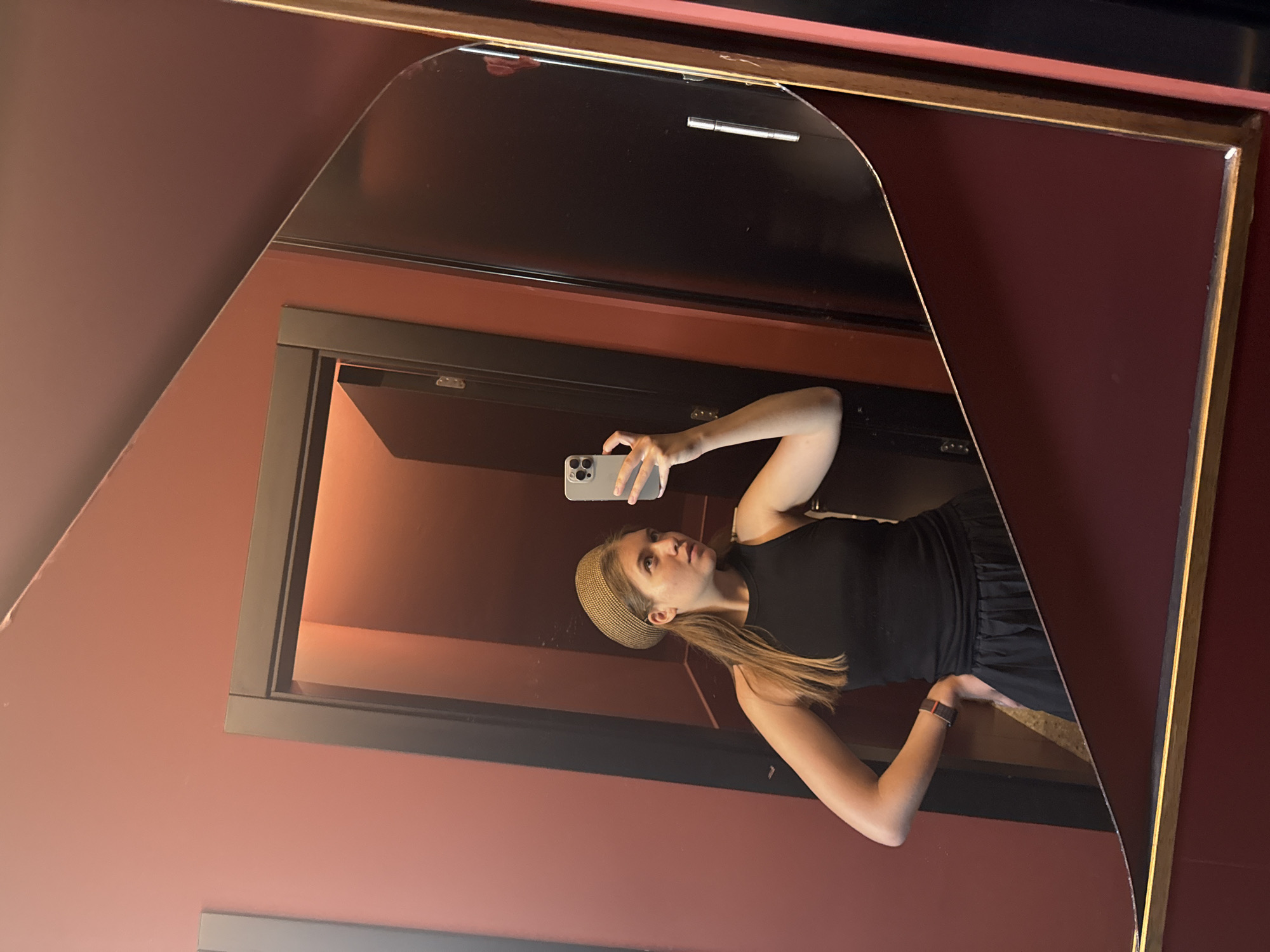 A woman is taking a selfie in front of a mirror on a wall. The mirror is framed with a golden border, which contains a brown, rectangular door. The woman is wearing a black sleeveless top, a brown headband, and an Apple watch. The wall is red. The location may be in Belgrade, Serbia.