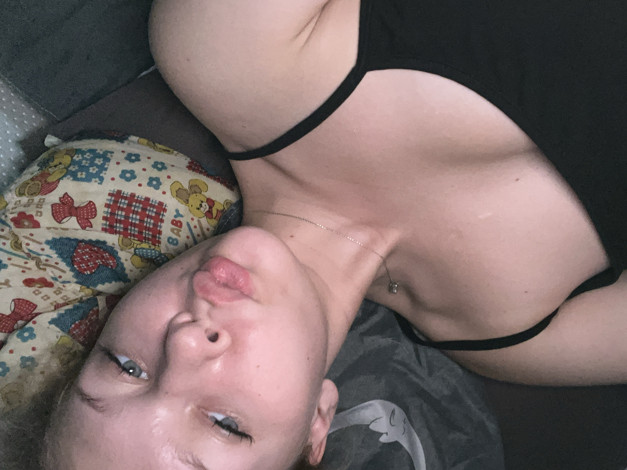 A close-up, high-angle, waist-up selfie shows a young woman with light skin and light brown hair, lying on her side on a bed. She is wearing a black camisole with thin straps. Her head is tilted and her lips are pursed, as if blowing a kiss. She is looking slightly off-camera to her left. Behind her is a pillow with a colorful pattern featuring cartoon animals and floral designs. The pillow appears to be a baby pillow. The background is dark and out of focus. The lighting is soft and appears to be artificial. There is no indication of the location being in Perm, Russia.