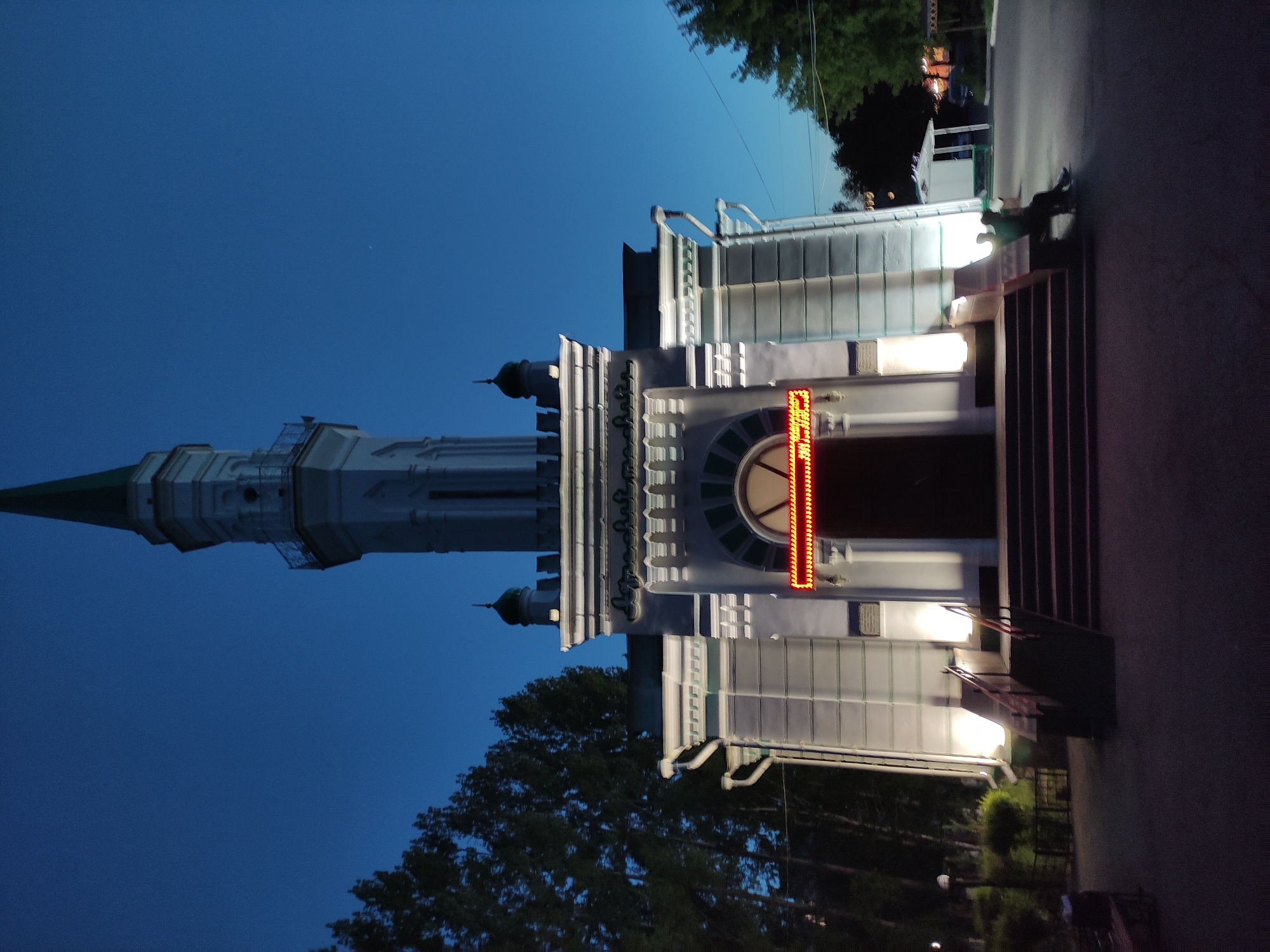 The image shows a building with a tall, white tower under a blue sky at night. The building appears to be in Pavlodar, Kazakhstan. The building's entrance is illuminated, and it has a display with lights. There are trees in the foreground and street lights in the background. Some people are visible in the shadows, sitting by the stairs.
