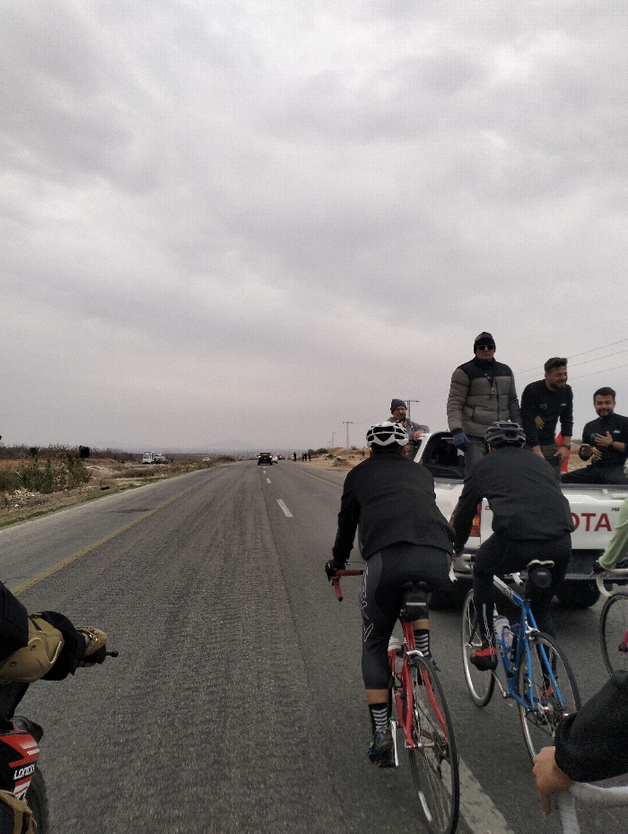 The photo depicts cyclists riding on a road with a support vehicle nearby. Several men stand in the back of a white Toyota pickup truck, some observing the cyclists. The sky is overcast. Other vehicles are visible on the road ahead. The cyclists are wearing helmets and riding gear, and the camera appears to be positioned from behind another cyclist or motorcycle.