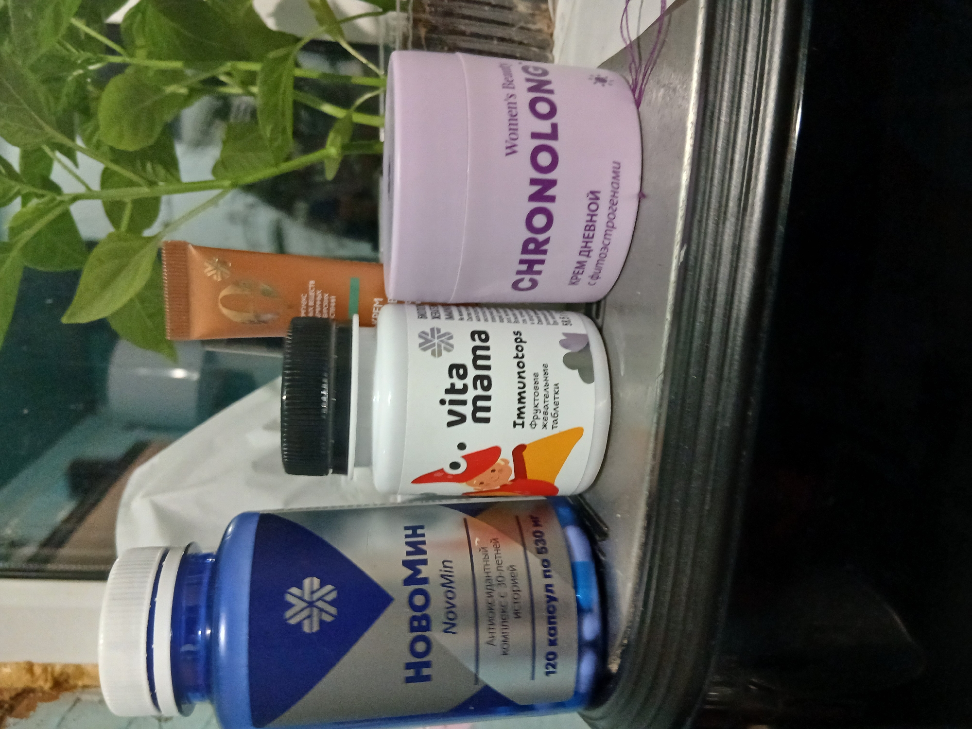 The image shows a collection of personal care products arranged on a surface, possibly a shelf or counter. From left to right, there is a blue bottle labeled "HOBOMИH NovoMin," a white bottle with a black cap labeled "vita mama Immunotops," and a lavender container labeled "CHRONOLONG." There is also an orange tube of what appears to be cream, as well as a green plant in the background. The presence of Cyrillic text on all the products suggests the location is within a Russian-speaking area. Based on the text on one of the products, the city is likely to be Ильинка, Russia.
