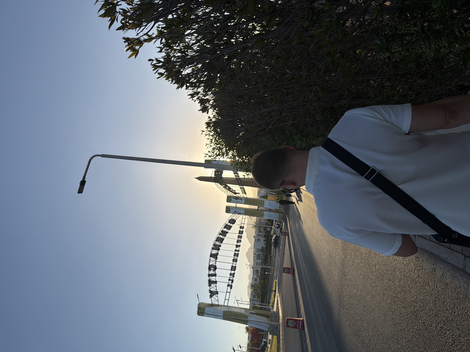An overhead shot captures a man walking away from the camera down a paved road in Sharm El Sheikh, Egypt. The man is wearing a white polo shirt and a black crossbody bag. The road is lined with trees on the right side of the image. In the background, there is an arched gateway with the words "Shopping Mall" visible. The sun is setting behind the gateway, casting a warm glow on the scene. A street lamp stands to the left of the man. The sky is a clear blue. There are a few cars and people in the distance.