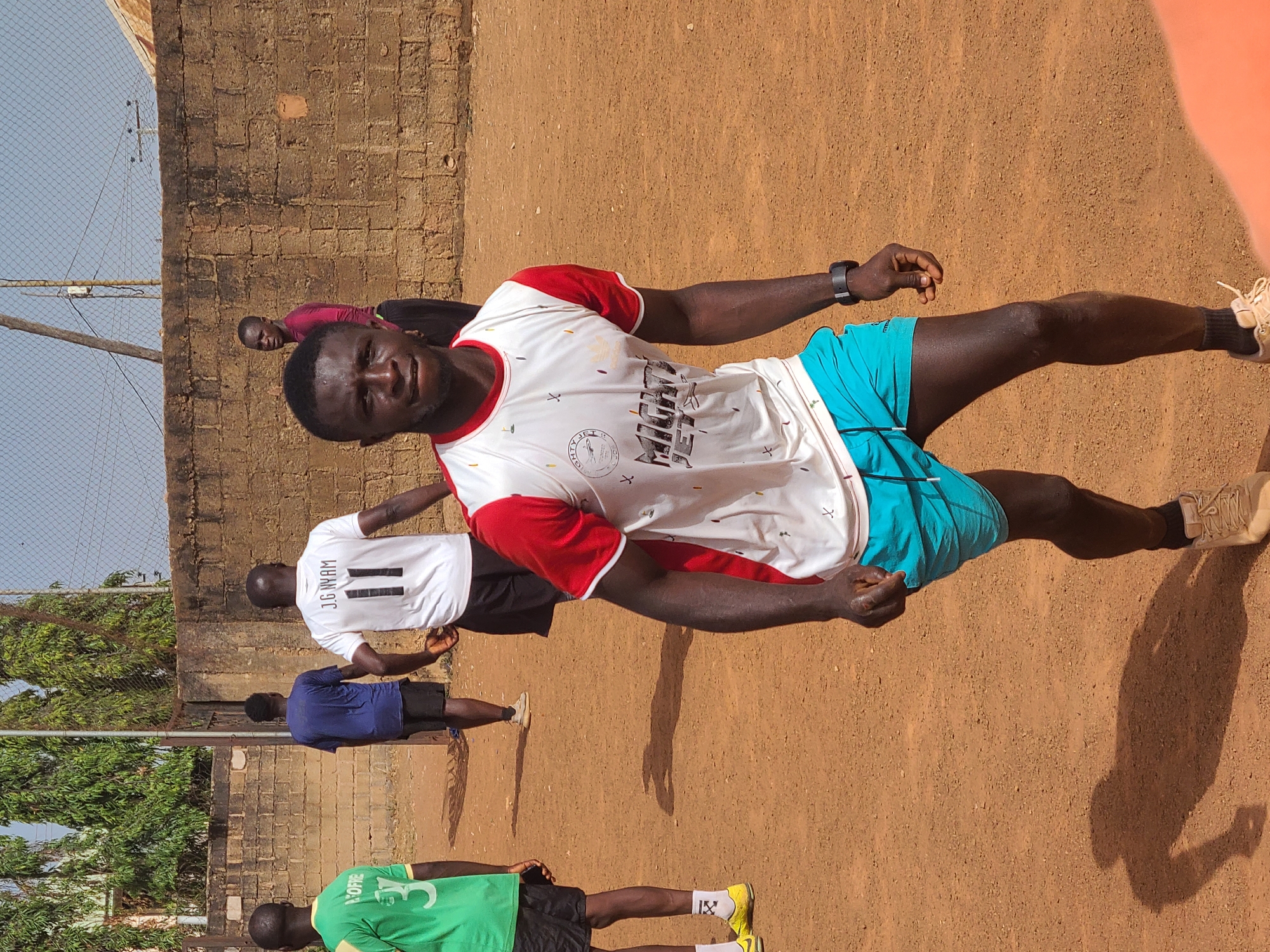 The image shows a group of young men standing on a dirt field, seemingly in Barakin Lerawi, Nigeria. The central figure is a man wearing a white t-shirt with red sleeves and blue shorts, looking directly at the camera. Behind him are other young men in various sports jerseys: one in a white jersey with "J.G NYAM" and the number 11 on the back, another in a blue shirt, and one in a green jersey. The background includes a brick wall, a chain-link fence, and sparse trees. The ground is dry and reddish-brown. The lighting suggests it is daytime.