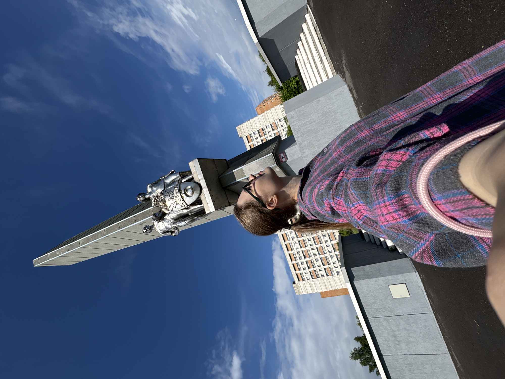 A low-angle, wide shot captures a woman in a plaid shirt looking up at a monument in Kirovo-Chepetsk, Russia. The monument, a tall, gray, obelisk-like structure, is topped with two metallic figures and a circular emblem. The woman, with her hair in a ponytail and wearing glasses, is positioned in the foreground, her upper body and face visible as she gazes upwards. Behind the monument, residential buildings and trees form the urban backdrop. The sky is a vibrant blue with scattered white clouds. The ground is paved and appears to be an outdoor public space.
