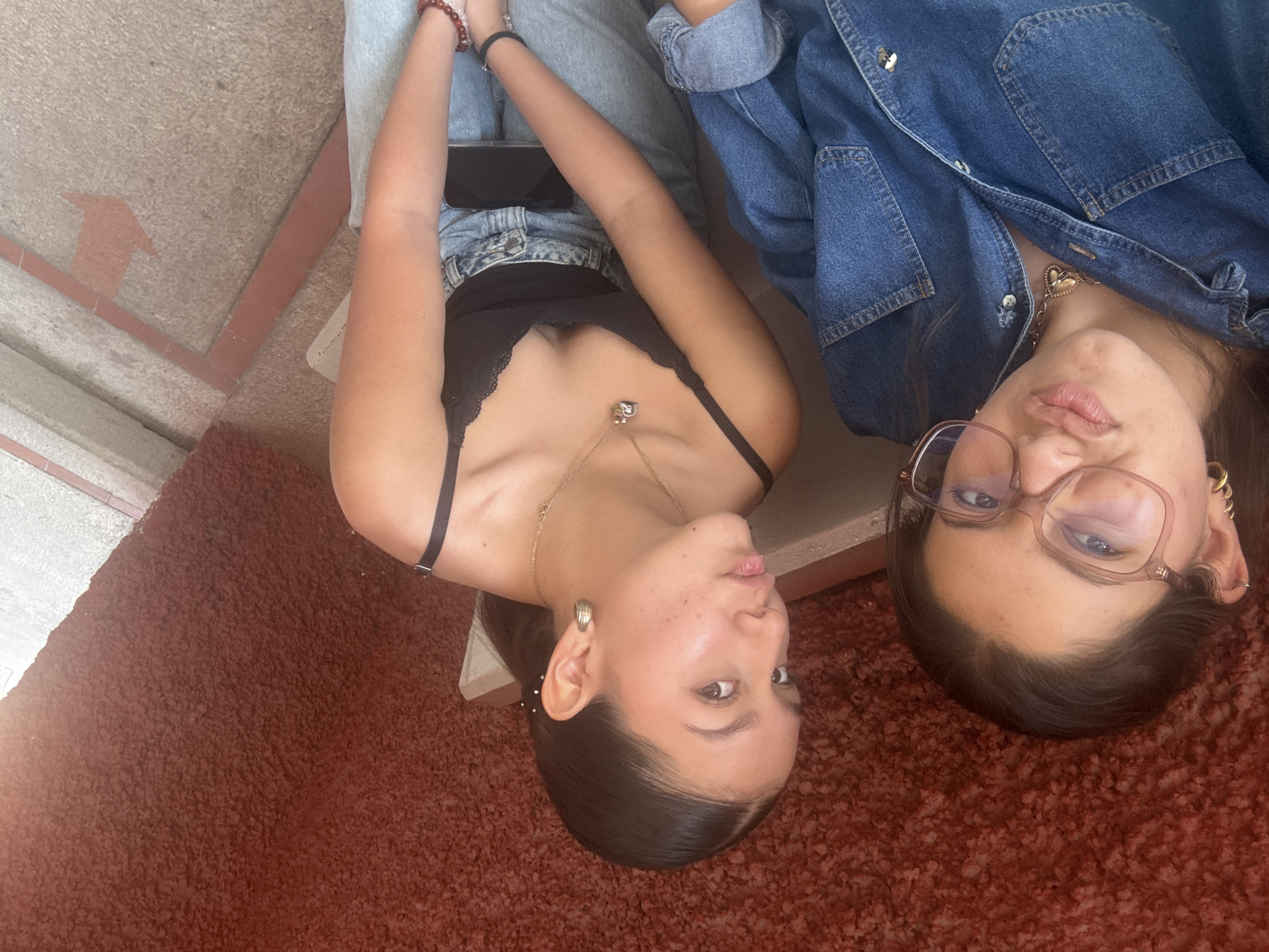 Two young women are lying on the floor, close to each other. The floor is covered in a textured, reddish-brown material. The woman on the left wears a black top with lace detailing, light-colored jeans, and a gold necklace. She has dark hair pulled back and is looking at the camera with a slight pout. The woman on the right wears glasses and a denim jacket, also with a necklace, and is making a similar facial expression. They are situated on a concrete step and wall area, near an arrow symbol painted on the concrete. This image was taken in San Andrés Cholula, Mexico.