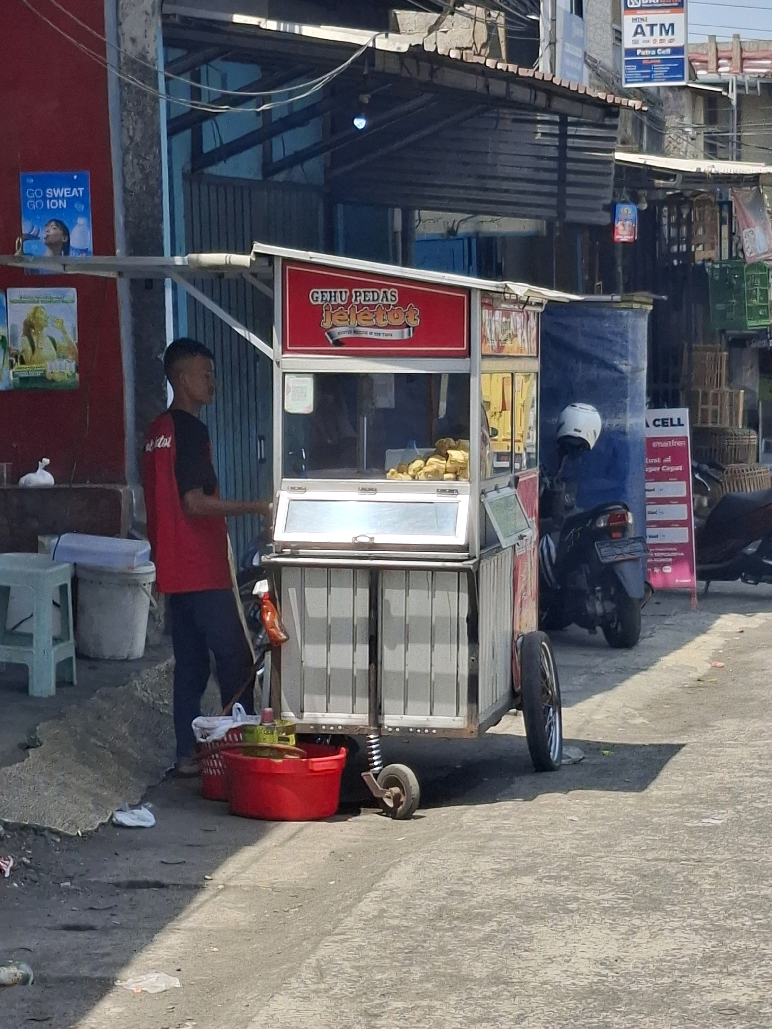 This image depicts a street scene in Cikahuripan, Indonesia, focusing on a food cart. A young man wearing a red t-shirt with black sleeves stands behind the food cart, which is primarily red and silver. The cart displays a sign with Indonesian text, reading "GEHU PEDAS JELETOT." Inside the cart, fried food items are visible. To the right of the cart, a motorcycle with a white helmet is parked, partially covered by a blue tarp. Several red plastic baskets and containers are on the ground near the food cart. In the background, there are buildings with awnings and various signs, including a "BRI" ATM sign. The ground is paved, with some debris present. The overall environment appears to be an outdoor market or street vendor area.