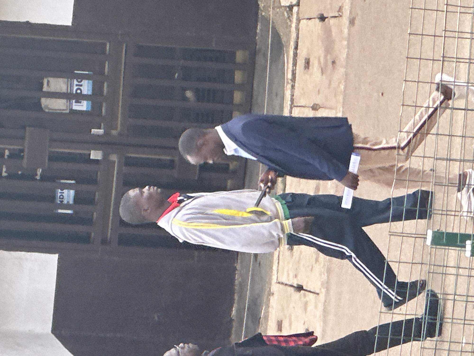 The image depicts an outdoor scene, likely in the city of Bukuru, Nigeria, with three individuals visible. In the foreground, a man wearing a dark blue suit and light blue shirt is walking from right to left, holding a white rectangular object in his left hand. He is also looking downwards. To his left, another man in a grey hooded jacket with yellow and green accents is walking in the same direction, his left hand is extended, holding a dark object, possibly a phone, and he is looking forward. To the far left, a third person, partially visible, is wearing a black garment with a red and black plaid pattern over their shoulder. They are facing left. The background features a dark building with barred windows and two signs with the text "POLICE" visible on them. The ground appears to be paved with dirt and gravel. A metal fence runs across the bottom of the image.