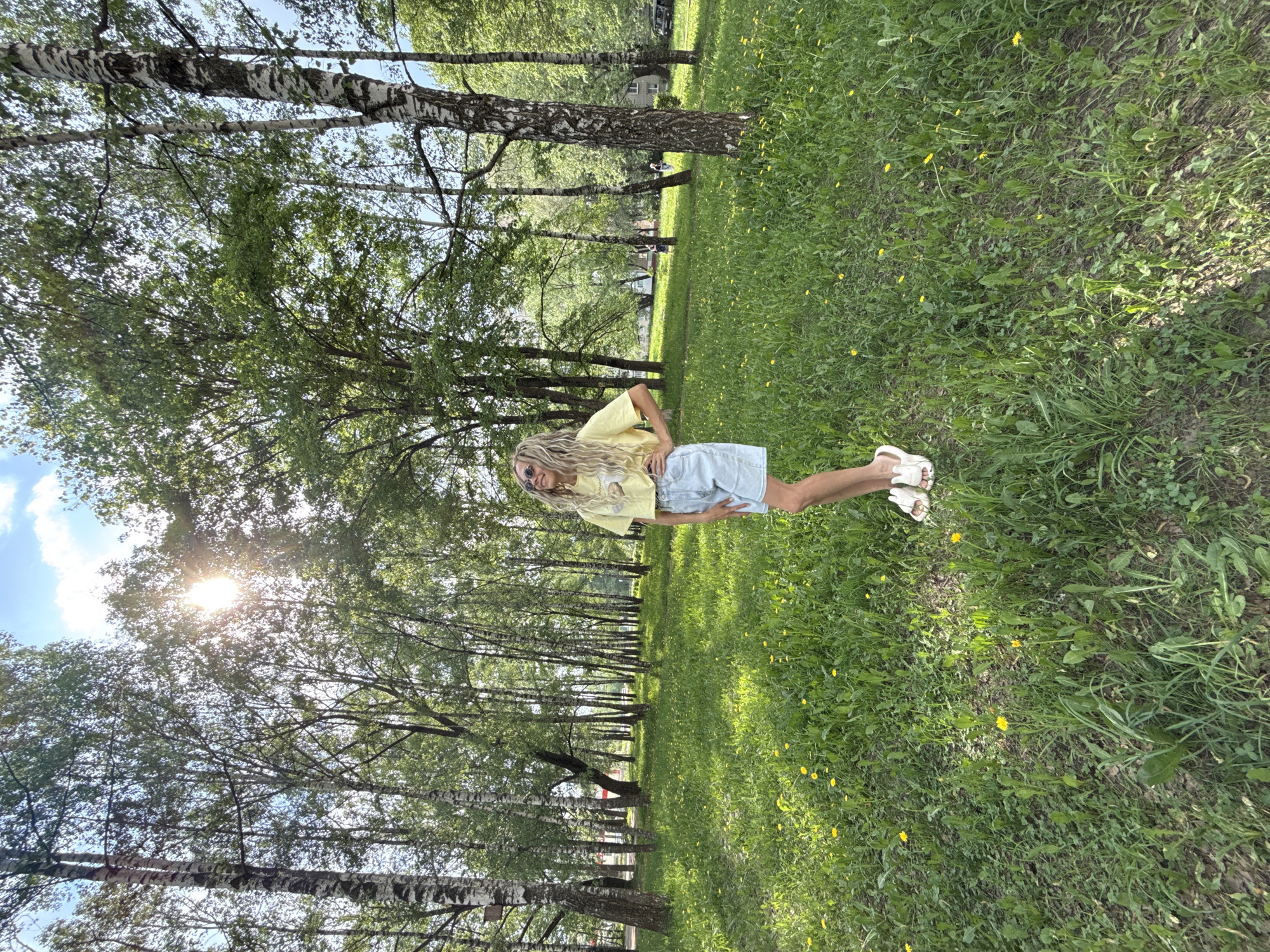 A blonde woman wearing a yellow shirt and light blue shorts stands in a park with birch trees in Rusanovo, Russia. She has her hands on her hips. The ground is covered in green grass and small yellow flowers. The sun shines through the trees.