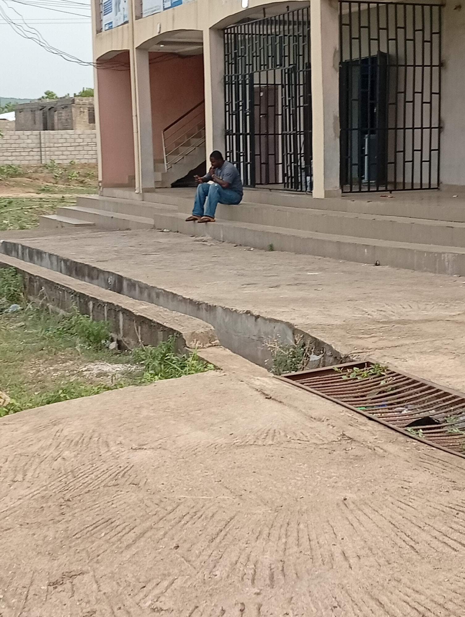 The image shows a man sitting on the steps of a building in Adankolo, Nigeria. The building has barred windows and a set of stairs leading to an upper level. The man is wearing a gray shirt and jeans. In front of the steps is a concrete area with a drainage system covered by a rusty grate. Vegetation grows in the ditch alongside the concrete area. In the background, there is a brick building under construction.