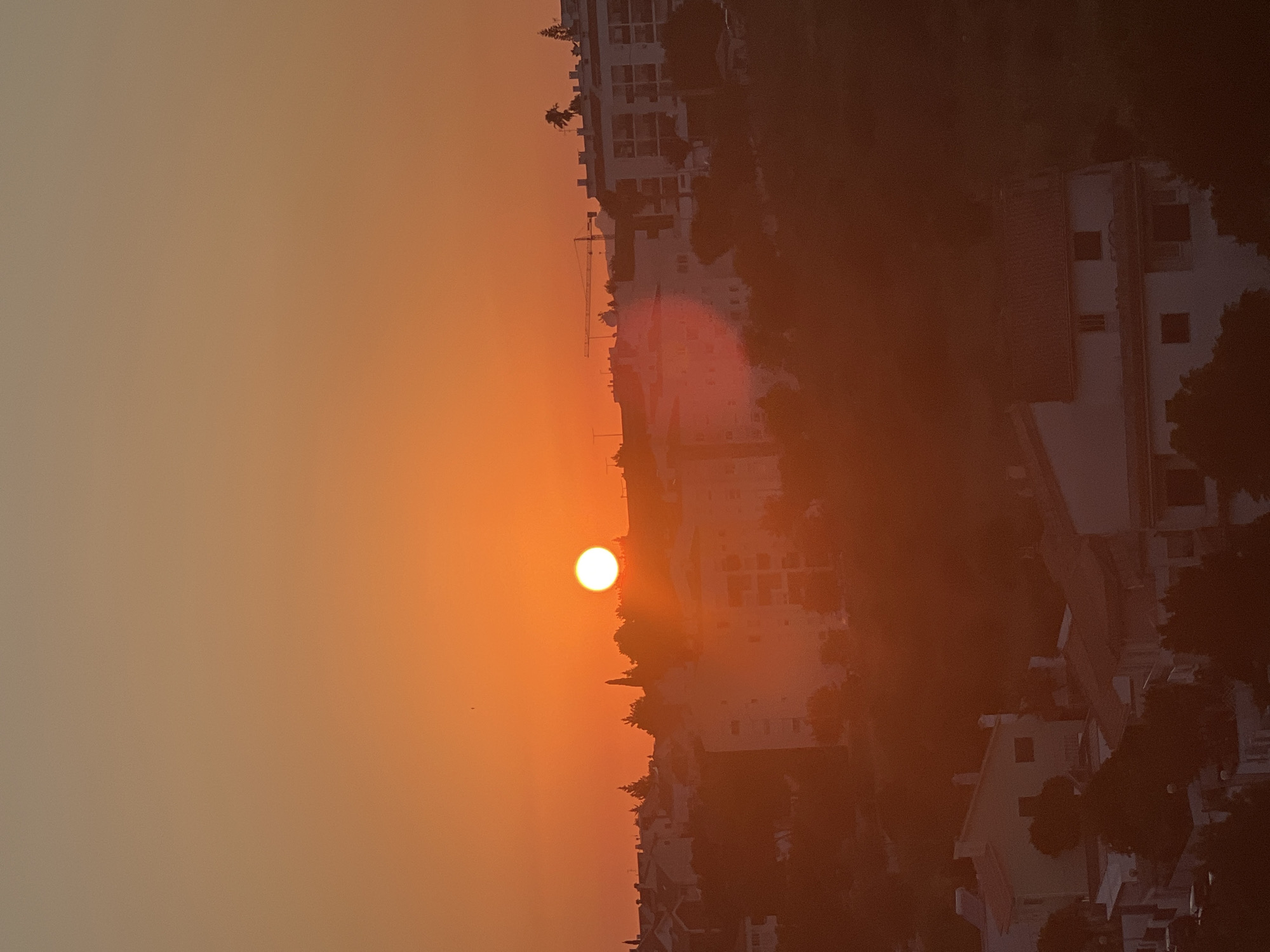 A wide shot captures a sunset over a city, likely Cascais, Portugal, as indicated by the architectural style of the buildings which are characteristic of the region. The sun, a bright orange orb, is prominently positioned in the sky, casting a warm, diffused glow across the scene. The sky transitions from a deep orange at the horizon to a lighter, creamy hue at the top of the frame. Silhouetted against the vibrant sky are buildings of varying heights, some appearing to be apartment complexes with balconies and windows, while others are smaller residential homes with tiled roofs. Trees and vegetation are scattered throughout the landscape, partially obscuring some of the structures. In the distance, a construction crane is visible, suggesting ongoing development in the area. The overall atmosphere is serene and tranquil, with the warm tones of the sunset creating a peaceful ambiance.