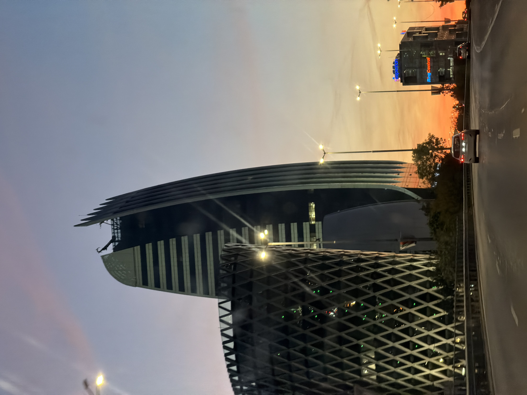 A low-angle view shows a building with a distinctive sail-like design in Sofia, Bulgaria. The building's structure is partially obscured by an adjacent structure with a diamond-patterned facade. A road runs alongside the building. Streetlights are visible. The sky displays a gradient from light blue to orange. A building with illuminated signs is in the background. Cars are visible on the road.