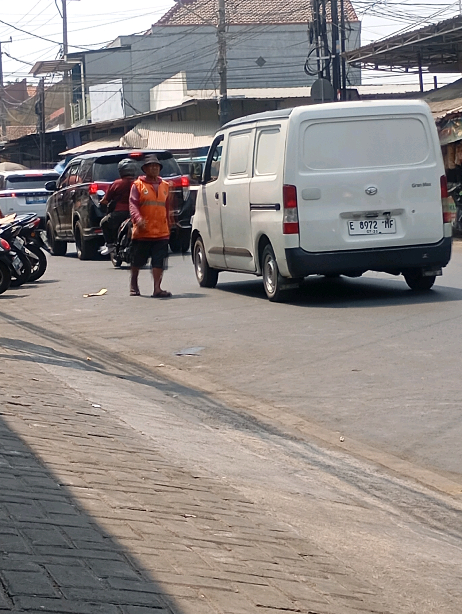 This is a horizontal, eye-level, medium shot taken on a sunny day in Sumber, Indonesia, capturing a street scene with traffic. In the foreground, a person wearing an orange vest and a hat walks on the road between a black SUV and a white van. The person is wearing dark shorts and appears to be walking away from the camera. Behind the person, a motorcycle is visible. The white van has a license plate that reads "E 8972 MF". In the background, there are buildings with tiled roofs and power lines crisscrossing the sky, indicating a typical urban or semi-urban setting. The road surface is asphalt, and the sidewalk to the left is paved with interlocking bricks. The overall atmosphere is that of a busy street with ongoing vehicular activity.
