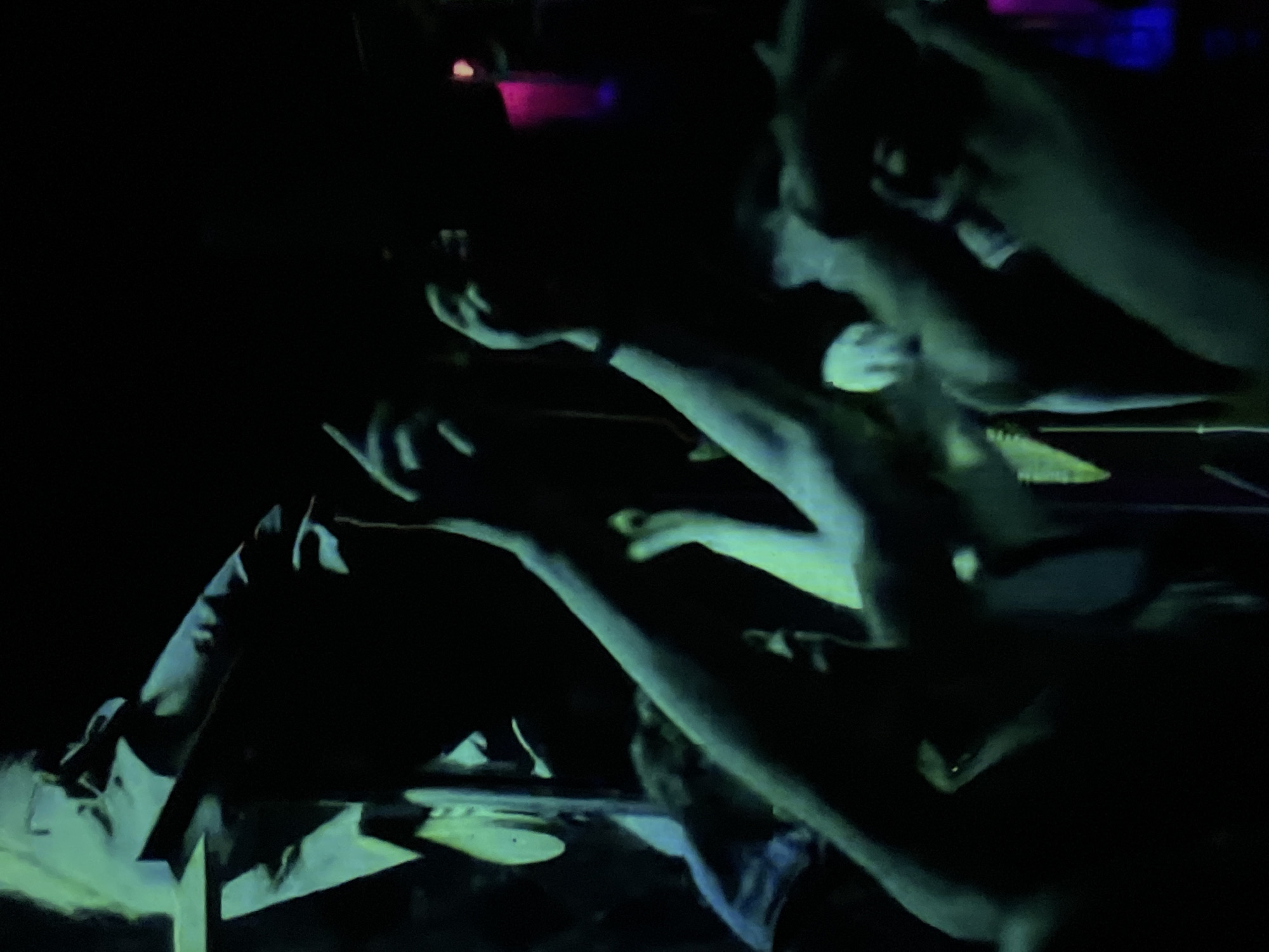 The image depicts a dimly lit scene, likely a concert or performance in an indoor venue. A crowd of people are visible, with their arms raised in the air. A person lies on what appears to be a platform or stage, with a dark jacket. The overall atmosphere is dark and active. There is insufficient information to ascertain a specific location within St. Petersburg, Russia.