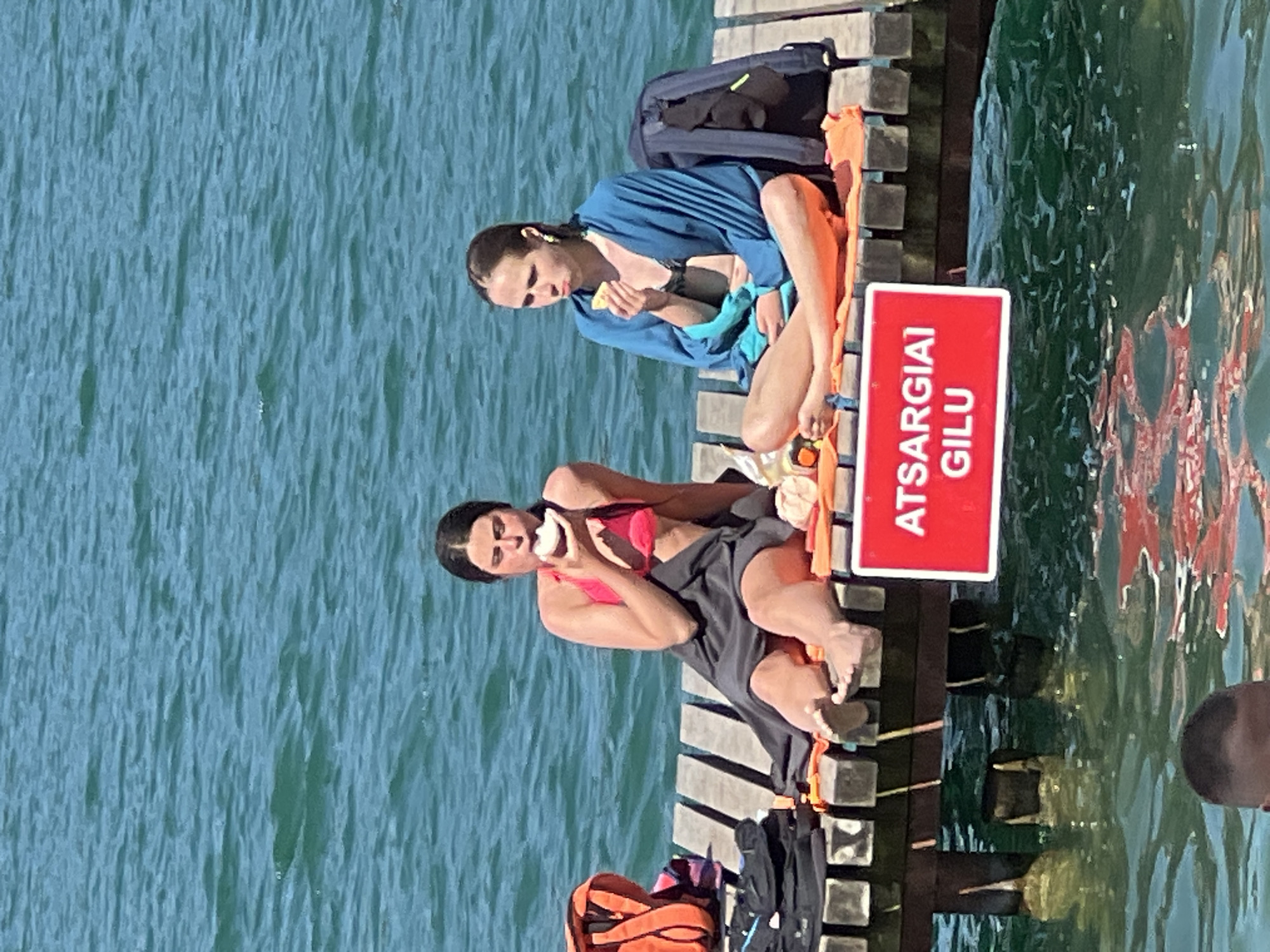 Two individuals are seated on a wooden pier on a sunny day. The pier extends over calm, turquoise water.
The subject in the foreground is a woman wearing a pink bikini top and a dark skirt. She is holding a piece of food and appears to be eating it. Her legs are crossed, and her feet are resting on the pier.
Behind her, another person, likely a woman, is seated with her legs outstretched. She is wearing a blue robe or cover-up over a swimsuit. She is holding a piece of food and looking down as if about to eat. A dark blue backpack is placed behind her on the pier, along with an orange towel.
A red sign with white lettering is attached to the side of the pier, facing the water. The text on the sign reads "ATSARGIAI GILU". This translates from Lithuanian to "CAUTION DEEP".
The water is clear and reflects the surroundings. The overall scene suggests a relaxed atmosphere, possibly a break from swimming or sunbathing. The time of day appears to be during daylight hours, indicated by the bright sunlight and clear visibility. The location appears to be a body of water suitable for swimming, with a pier for access. The presence of swimwear and towels suggests a recreational activity.