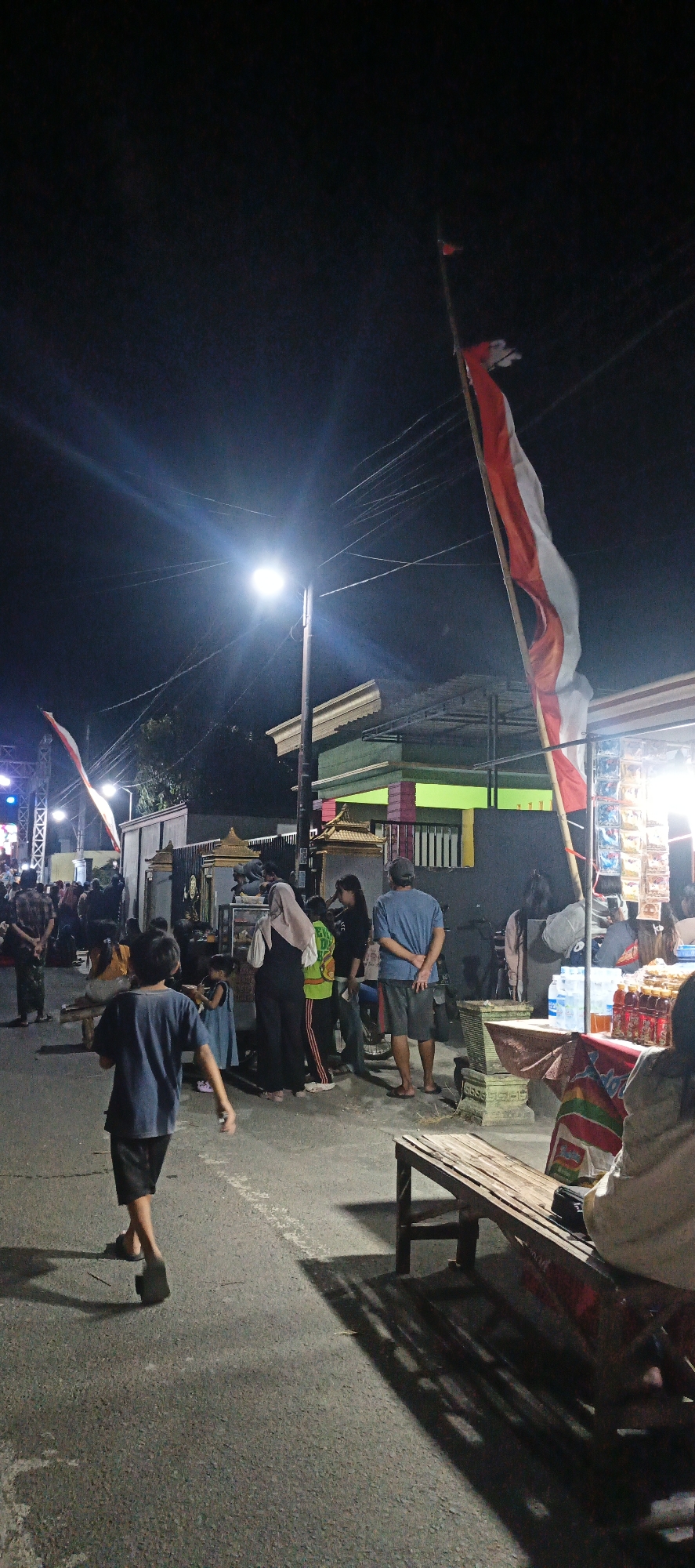 A street scene at night shows a crowd of people gathered around food stalls and vendors. An Indonesian flag is prominently displayed on a pole, waving in the breeze. Streetlights illuminate the area, casting long shadows. In the foreground, a young boy in a blue shirt and black shorts walks away from the camera. Several adults are seen standing and interacting at the stalls, some appearing to be making purchases. The background reveals buildings and structures typical of a residential or commercial area. The overall atmosphere suggests a lively night market or local gathering.