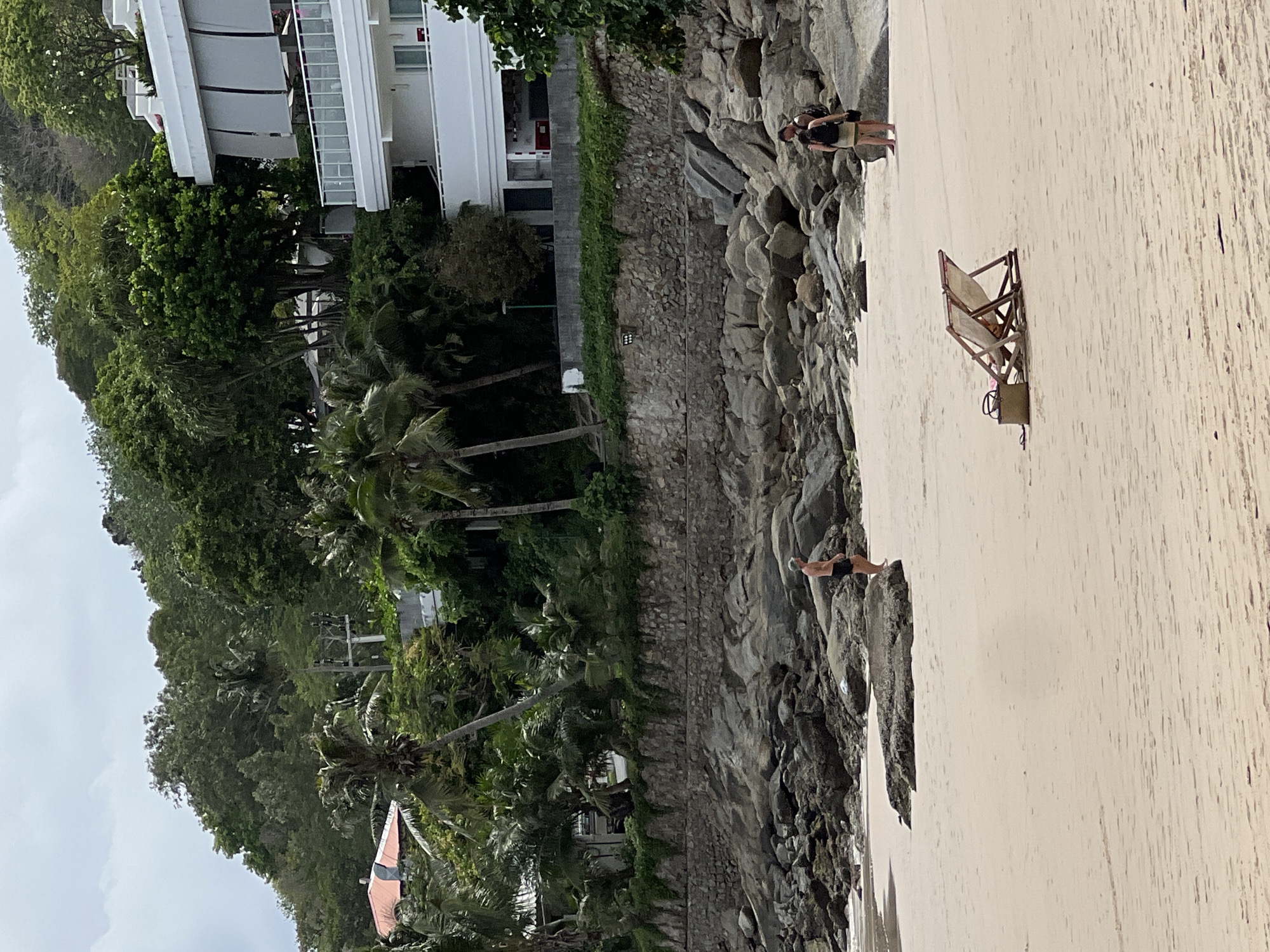 A vertically-oriented outdoor scene depicts a beach in Unknown, Unknown. Dominating the left is dense vegetation, including palm trees, flanking a multi-story white building. A stone wall separates the vegetation from the beach. On the beach, rocks line the edge closest to the vegetation. A beach chair with accompanying equipment sits on the sand. Three individuals are visible. One person is standing on the beach near the rocks, and a couple is standing further down the beach near the rocks.