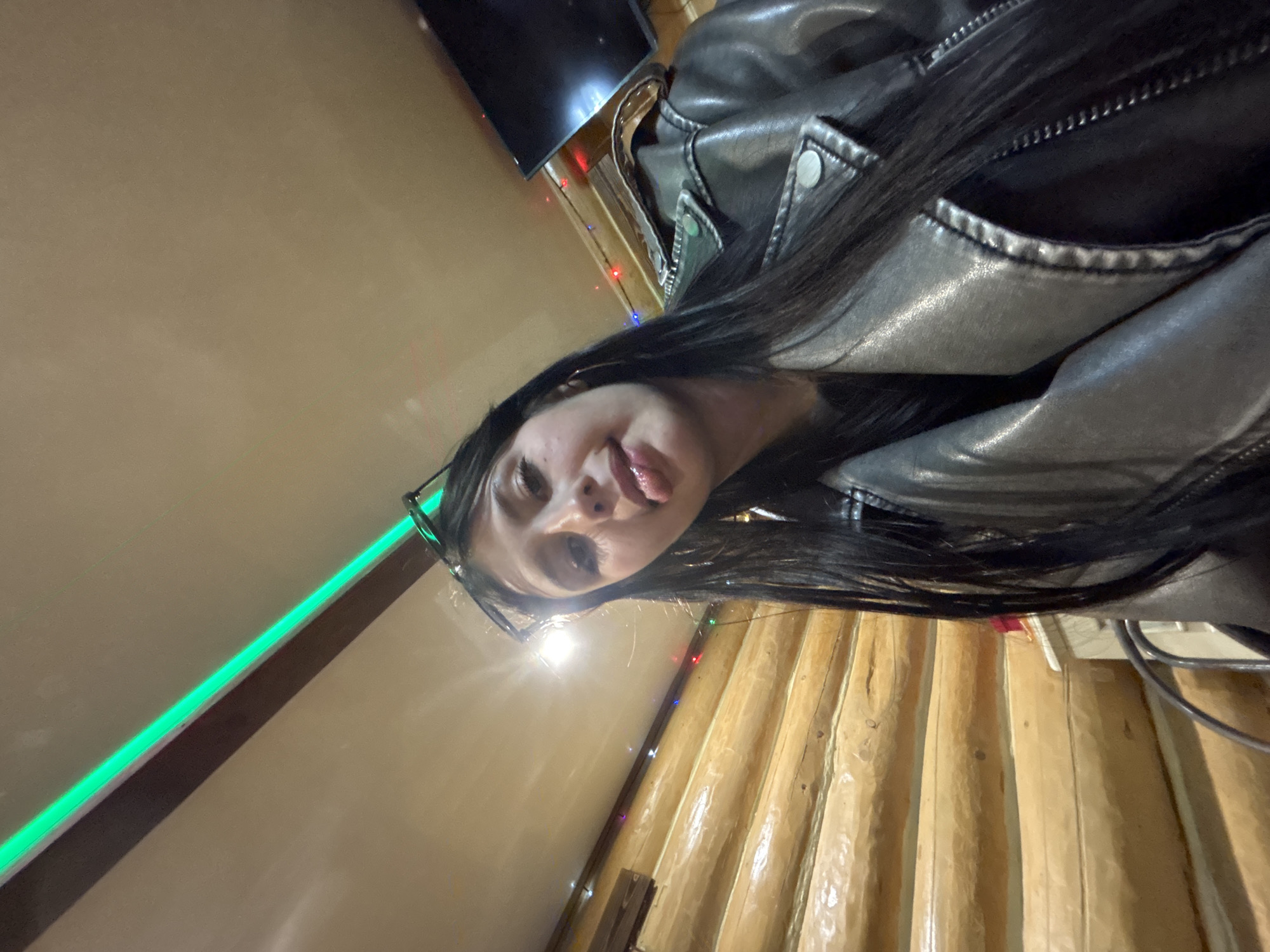 A selfie-style image taken in a dimly lit room in Chaikovsky, Russia. A young woman with long, dark hair is wearing a black leather jacket and glasses, and sticking her tongue out. The background consists of a light brown wall on the left with a green LED strip. A wooden wall is on the right, decorated with small colored lights. A television is in the upper left corner of the frame. Electrical cords are visible in the lower right.