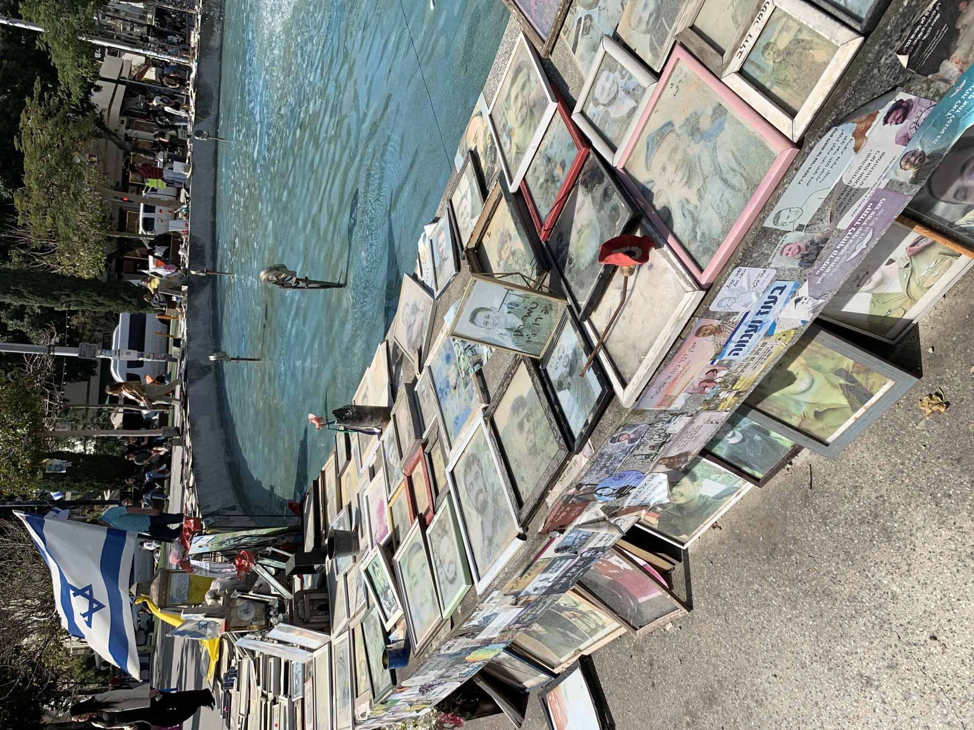 The image shows a memorial display along the edge of a water feature, likely a fountain, in Tel Aviv, Israel. The display consists of numerous framed photographs, mostly portraits, placed on the pavement. There are also printed materials with Hebrew text. An Israeli flag is prominently displayed to the left of the framed pictures, and artificial red poppies are interspersed among the pictures. Some people are standing in the background. The location is a public space in Tel Aviv.