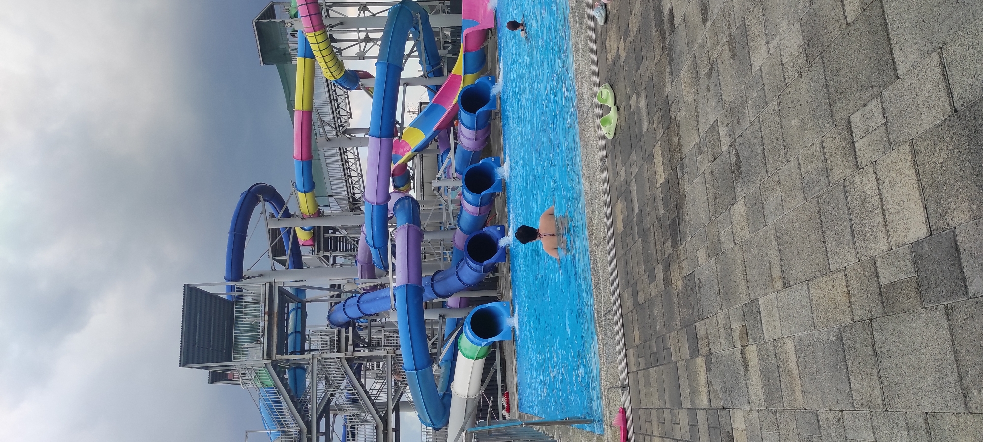 This media file depicts a scene at a water park in Tulskiy, Russia. The primary subjects are several colorful water slides, a swimming pool, and a few people. The water slides are a prominent feature, with various tubes in shades of blue, purple, pink, yellow, and green, constructed from metal and plastic. They are supported by a complex metal structure.
The swimming pool is filled with bright blue water, and the surrounding area is paved with grey, rectangular tiles. One person is visible in the pool, with their back to the camera, appearing to be wading. Another person is seen in the distance in the pool, and a third person is partially visible at the edge of the pool. There are also a pair of green flip-flops on the tiled ground.
The activity taking place is recreation at a water park, with the slides and pool suggesting a day of fun. The sky is cloudy, indicating possibly overcast weather. The time of day is not explicitly clear but appears to be daytime. There is no visible text within the image. The overall mood suggested is one of leisure and enjoyment, typical of a water park environment.