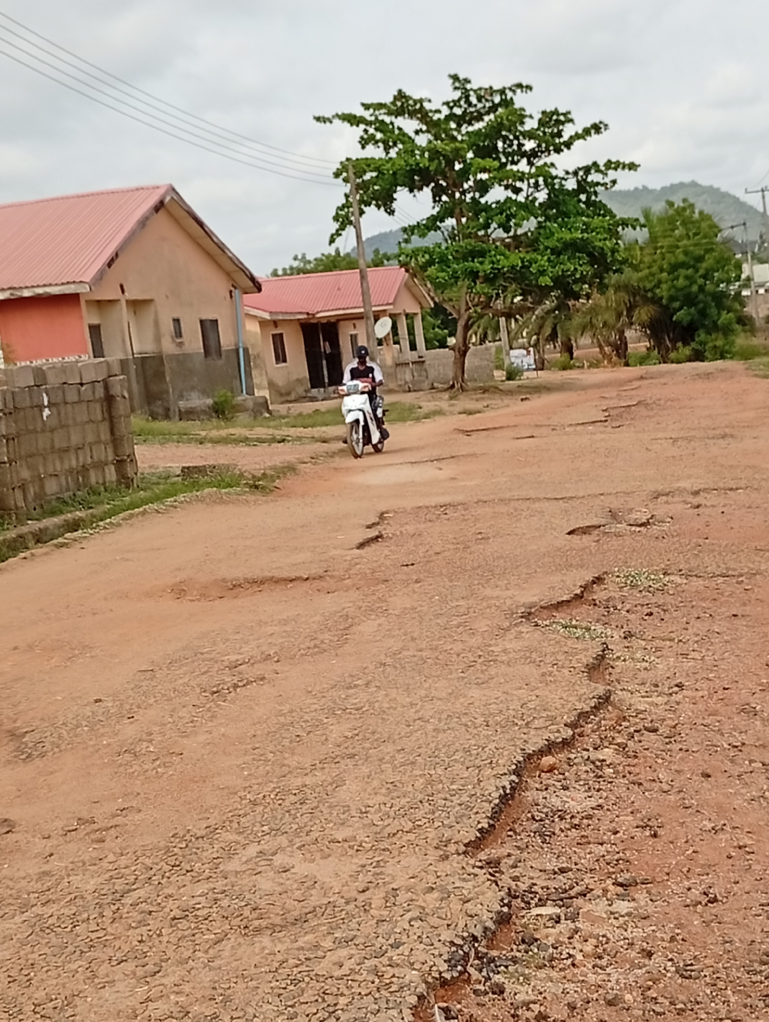 The image depicts a street in Adankolo, Nigeria. A motorcyclist is driving on a dirt road that is severely cracked and damaged. There are buildings with red roofs on either side of the road. Trees are visible, and there is a mountain in the background.