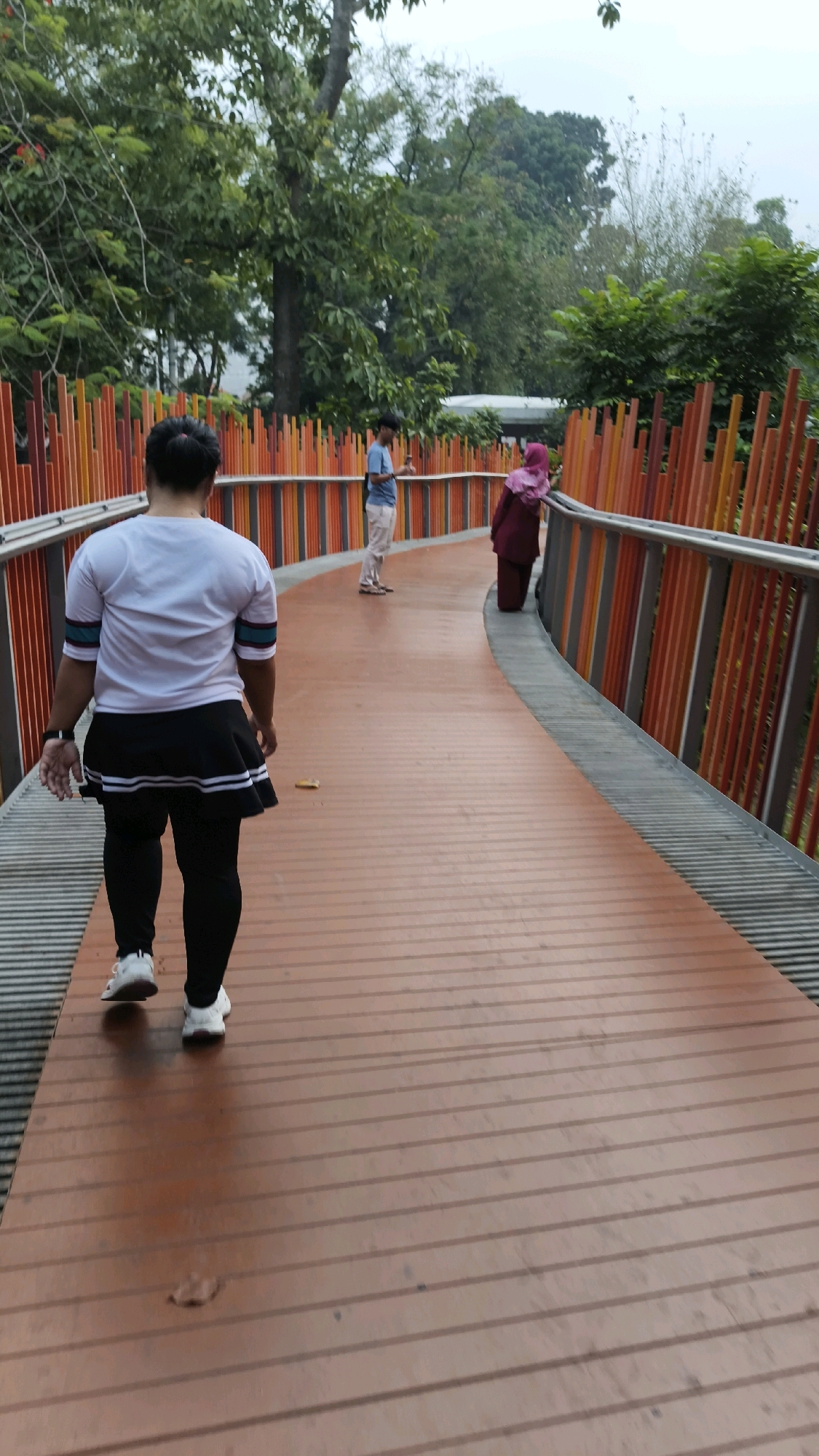 The image shows a curved pedestrian walkway with a wooden surface and a railing composed of vertical orange and red poles. In the foreground, a person wearing a white shirt, black skirt, black leggings, and white sneakers walks away from the camera. Further down the walkway, a person in a blue shirt and light-colored pants stands, and behind them, a person wearing a dark maroon hijab and dress looks out at the surroundings. The walkway is surrounded by lush green trees and foliage, suggesting an outdoor, possibly park-like setting. The overall lighting indicates an overcast day. This scene is located at the Skywalk in Kebayoran Baru, South Jakarta, Indonesia.