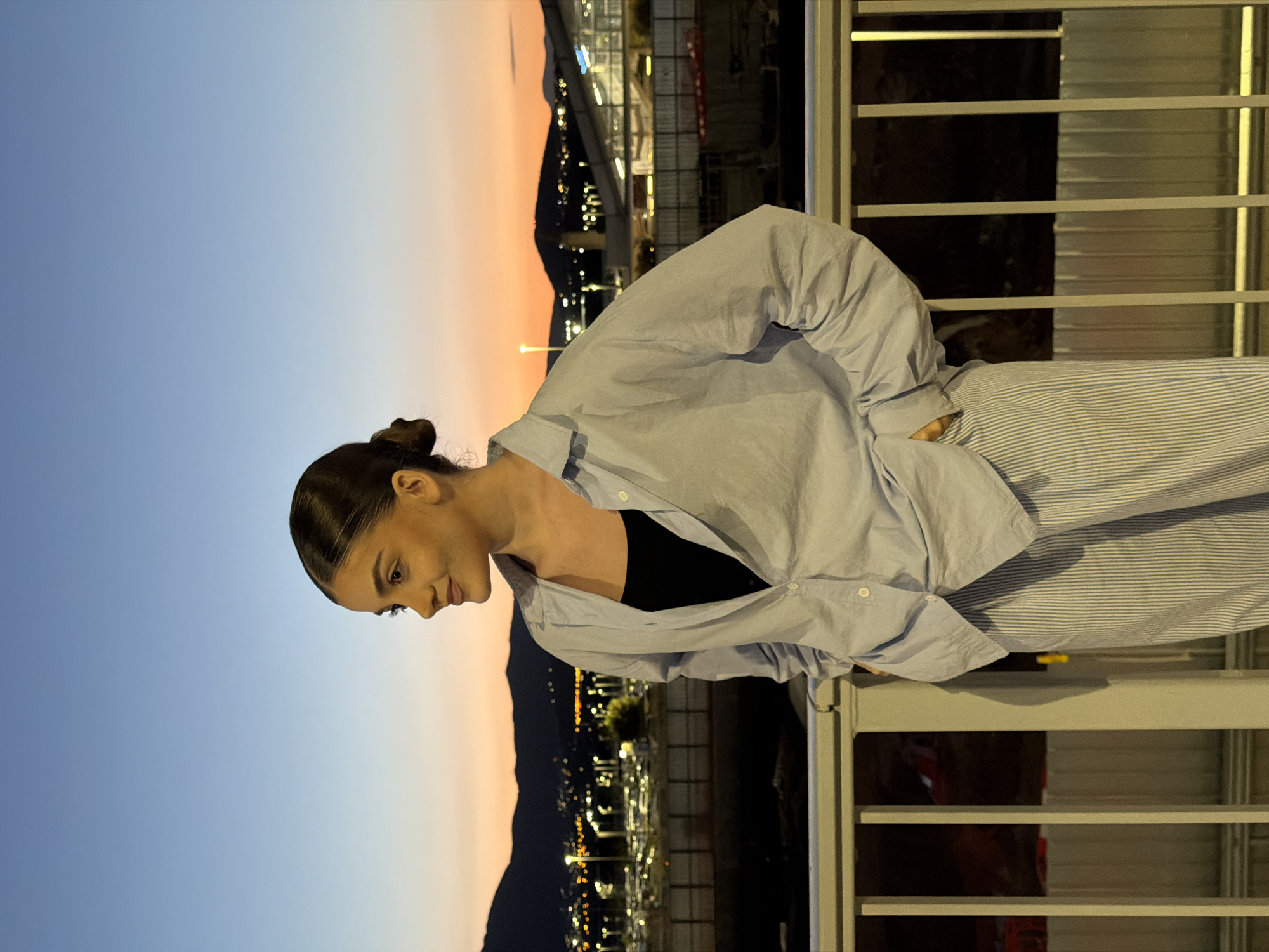 A woman stands on a balcony overlooking a city, presumably in Spata-Loutsa Municipal Unit, Greece, as indicated by the landscape and urban lighting in the distance. The time of day appears to be dusk or early evening, with a gradient of blue and orange in the sky. The woman has her hair pulled back in a bun and wears a black top under a light blue button-down shirt and striped pants. She leans slightly forward, looking down, possibly at the city below. The balcony has a metal railing with horizontal bars.