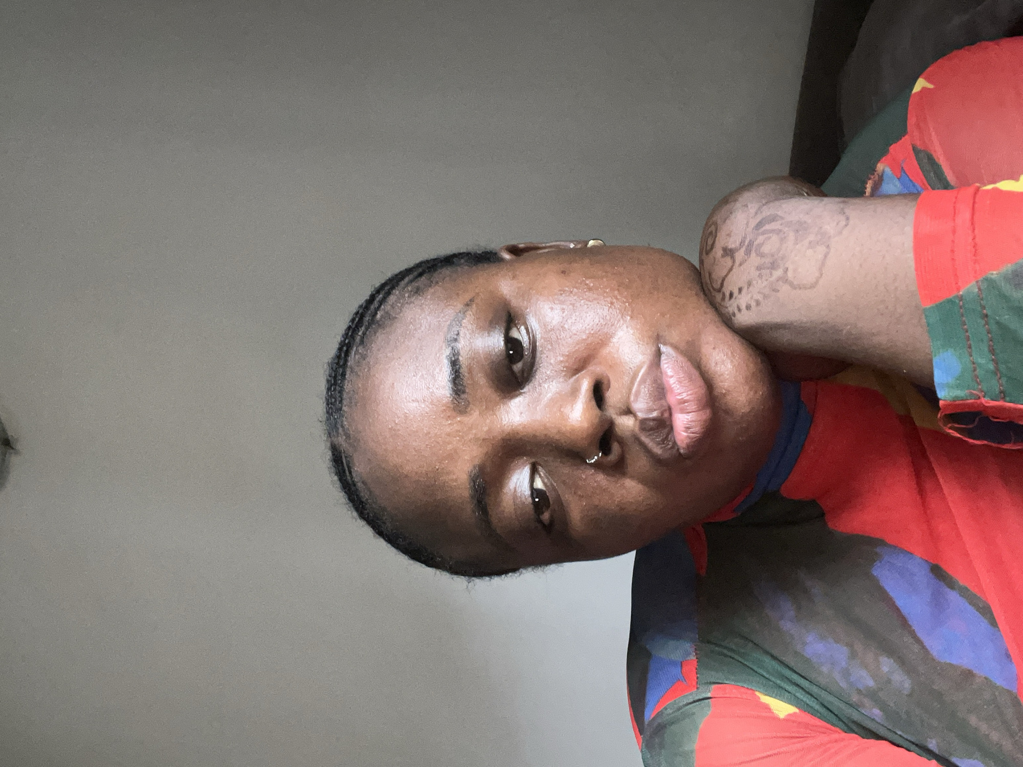 A close-up, angled portrait shows a person with dark skin and short braids, wearing a colorful shirt, possibly a t-shirt, with a design that includes red, blue, and green. The person has a nose ring in their left nostril and what appears to be a temporary tattoo on their neck. The background is a plain gray wall. There are no elements visible that suggest a specific location or interaction within Abuja, Nigeria.
