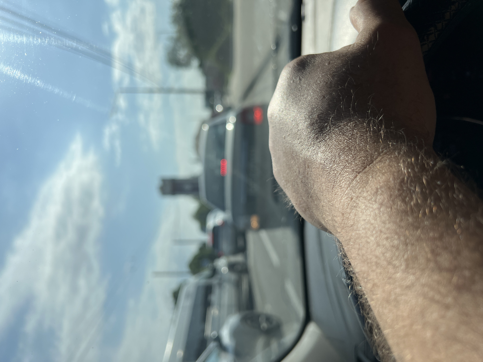 The image shows a view from inside a car, likely in traffic. A fair-skinned hand is gripping the steering wheel. Outside, through the windshield, there's a line of cars, including a silver minivan with its brake lights on. A tower is visible in the background. Based on visual elements, the image does not offer definitive clues to confirm this location.
