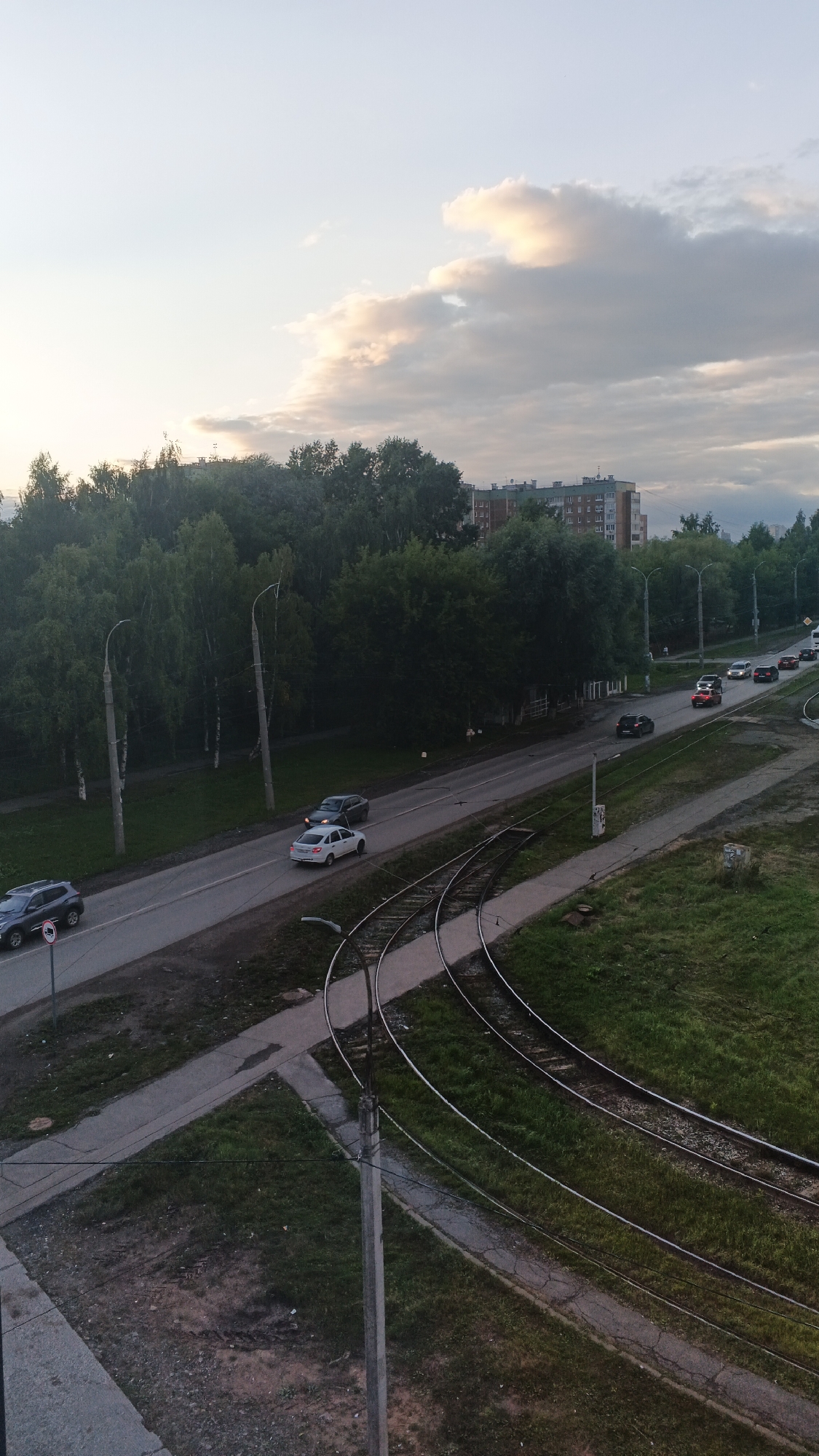 The image depicts an outdoor scene in Izhevsk, Russia, likely during the late afternoon or early evening. The sky is partly cloudy with soft, diffused light, suggesting a mild weather condition. The primary subjects are vehicles on a road and tram tracks running alongside it.
On the road, a line of cars is visible. A dark SUV is in the foreground on the left, followed by a white sedan and then a dark sedan. Further down the road, several other vehicles are seen, including a red car and a few other indistinct cars in the distance. The tram tracks curve through the scene, bordered by grassy areas and a paved path.
The setting appears to be a suburban or semi-urban area, with a dense line of deciduous trees forming a backdrop. Beyond the trees, a multi-story apartment building is visible, its windows reflecting the sky. The presence of streetlights along the road and the tram tracks suggests it is a developed area.
There are no people prominently visible in the image. The overall mood is quiet and uneventful, conveying a sense of everyday urban life. No visible text is present in the image. The composition and lighting suggest a natural, unposed photograph.