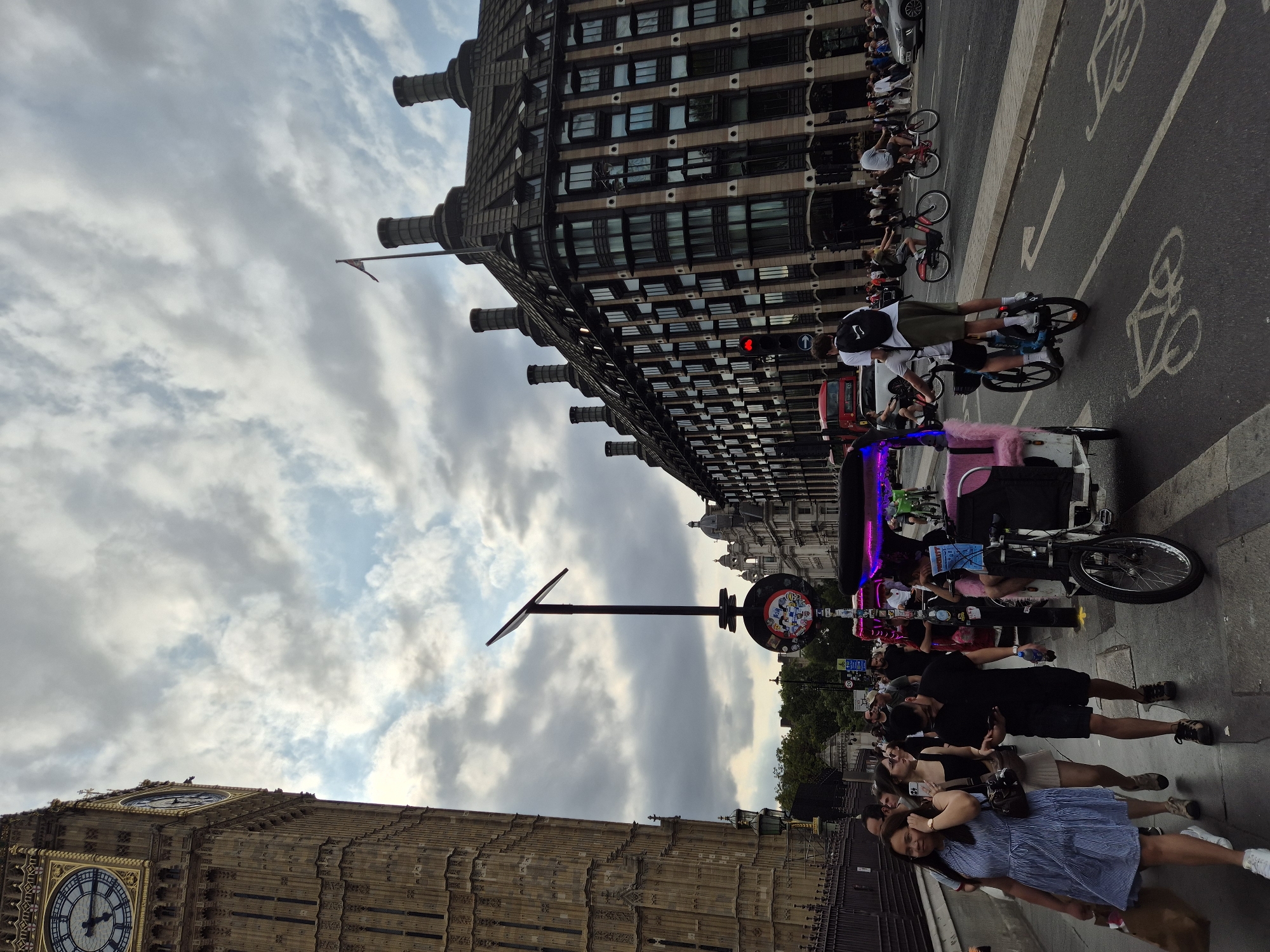 The image depicts a street scene in London, United Kingdom, with the iconic Big Ben clock tower prominently visible on the left. In the foreground, a group of pedestrians walks along the sidewalk. A woman in a blue and white striped dress is in the lead, carrying a shopping bag. Behind her, another woman in a black top and skirt walks with a phone in her hand, and a man in a black polo shirt and shorts looks at his phone. A pink pedi-cab with neon lights is parked on the right side of the sidewalk.
In the background, a large, imposing building with numerous windows and chimneys lines the street. Traffic, including a red double-decker bus and several cars, moves along the road. Two individuals are riding a bicycle together on the street, with one standing on the shoulders of the other. The sky is overcast with dramatic clouds, suggesting a day with variable weather. The overall scene captures a bustling urban environment with a mix of historical architecture and modern city life.