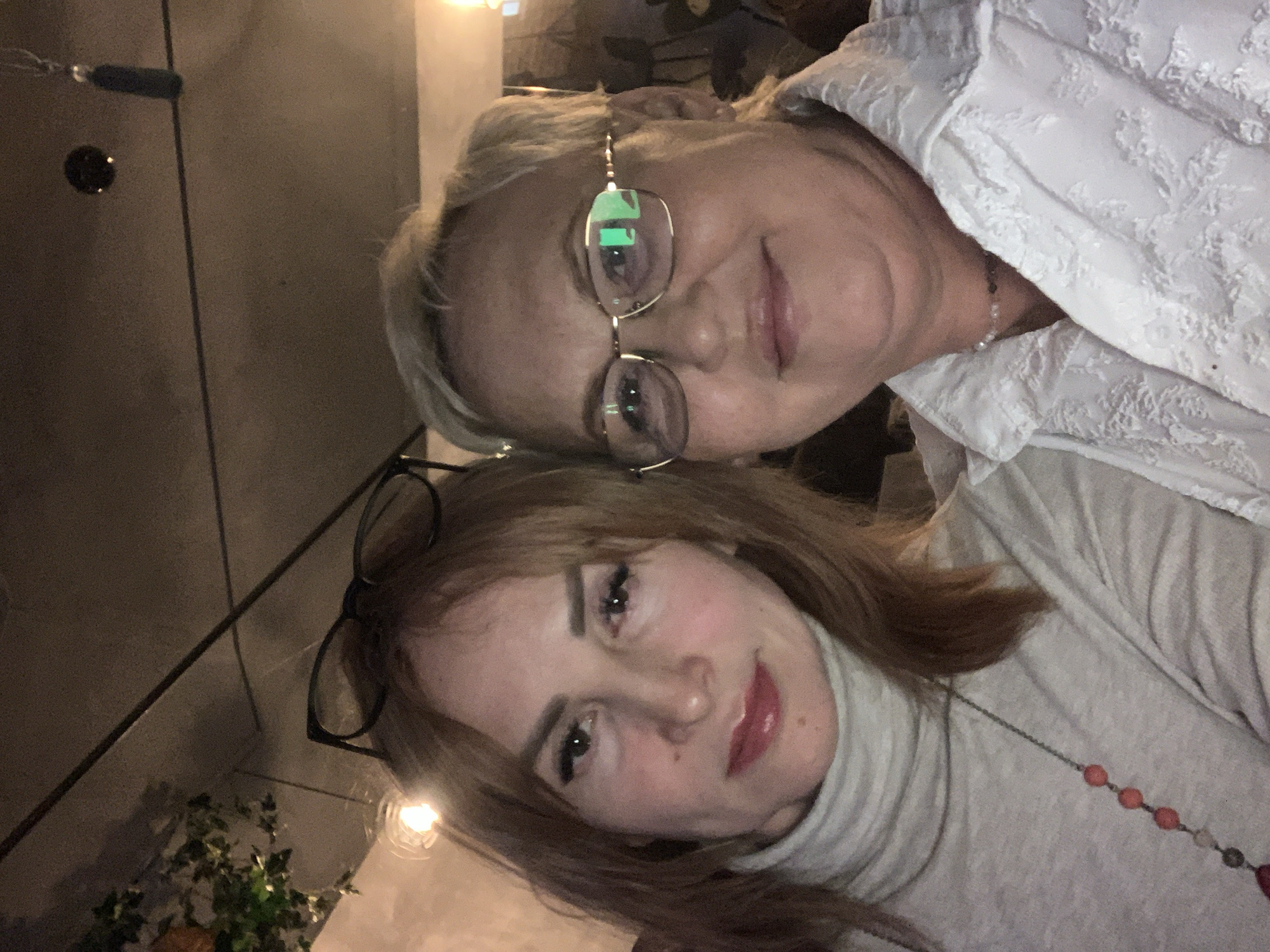The image captures two adult women posing closely together in an indoor setting, located in Chaikovsky, Russia.
The woman on the left has shoulder-length light brown hair with bangs, and dark-rimmed glasses are resting on top of her head. She is wearing a light beige or off-white turtleneck sweater and a long necklace with spherical red and translucent beads. Her facial features include defined eyebrows, eyeliner, and glossy lips, and she displays a slight smile while looking directly at the camera.
The woman on the right has short, light blonde hair and is wearing gold-framed rectangular prescription glasses, which show prominent reflections of light. She is dressed in a white collared shirt with a subtle embossed or textured floral pattern and a thin, multi-beaded choker necklace. She is smiling broadly and also looking towards the camera.
The background is dimly lit with warm artificial light. Above the woman on the left, a bright light source is visible. The ceiling appears dark, and on the upper left, a section of artificial green foliage or vines is discernible. On the upper right, a dark tassel-like decoration hangs from the ceiling.