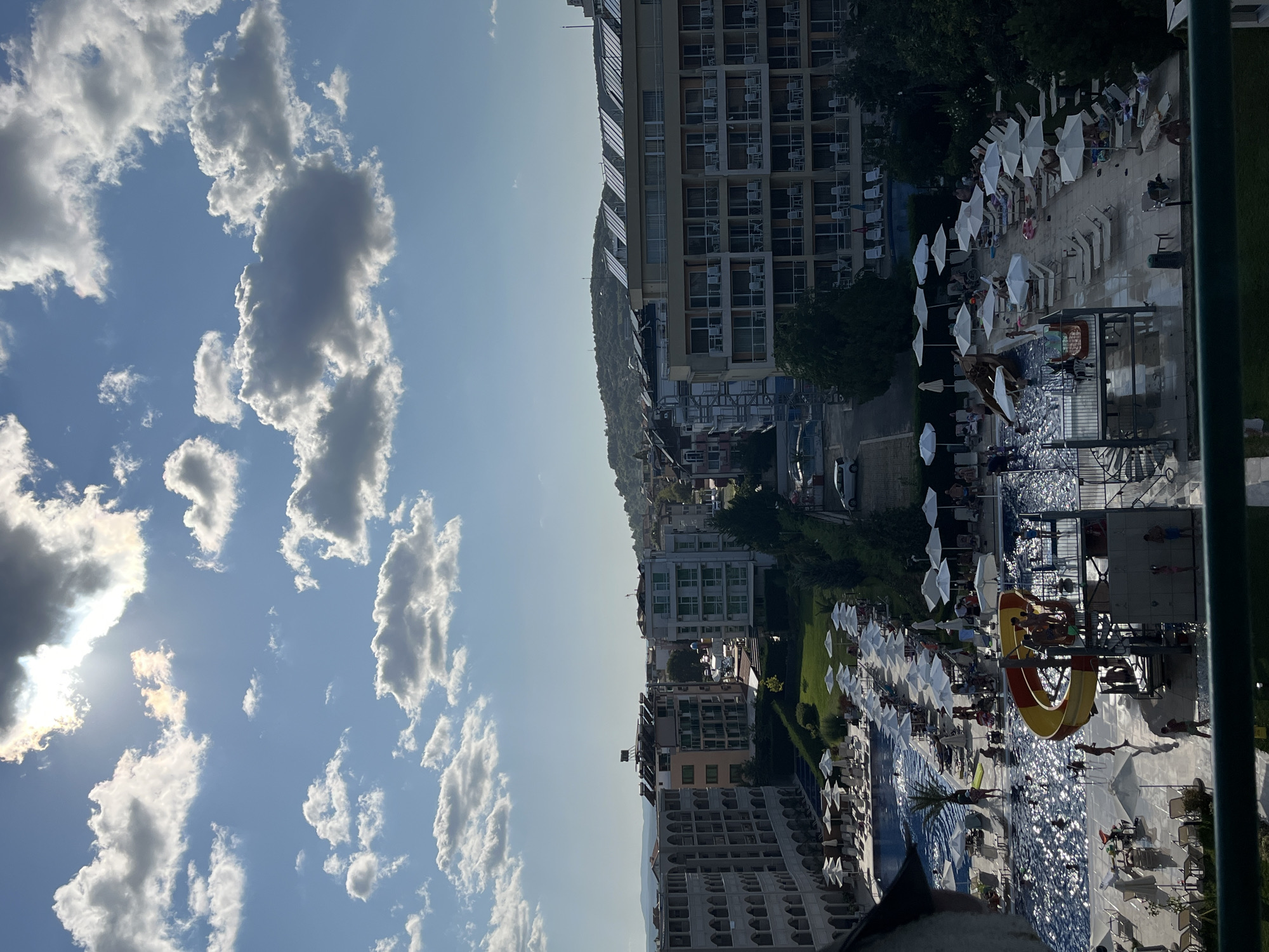 The image depicts a resort complex in Primorsko, Bulgaria, on a sunny day with scattered clouds. In the foreground, a large swimming pool with a water slide is visible, with several people swimming or relaxing by the poolside. Numerous lounge chairs and umbrellas are set up around the pool area, many of which are occupied.
Beyond the pool, rows of resort buildings with multiple stories are arranged. These buildings feature balconies and windows, suggesting hotel rooms or apartments. In the background, a lush, green hillside with some solar panels is visible, and the sky above is a clear blue with fluffy white clouds, with the sun shining brightly. The overall scene conveys a sense of vacation and leisure in a coastal resort town.