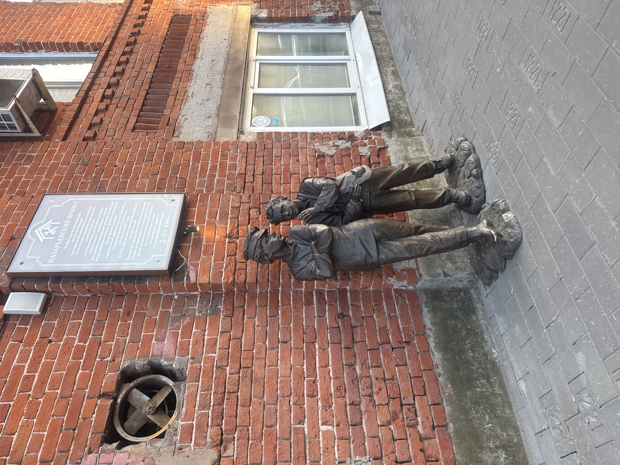 This image depicts a brick building with a plaque, window, and a ventilation fan. Adjacent to the building on a stone paved surface are two bronze sculptures of men lying down.
Location: It is located in Nizhny Novgorod, Russia. The plaque indicates that this is the location of the former Nazaryevsky Shelter.
Objects/Subjects:
- Two Bronze Sculptures: Two bronze sculptures of men, dressed in casual attire are lying down next to each other.
- Brick Building: A red brick building with a plaque bearing text in Russian.
- Window: A white-framed window on the second story of the building.
- Ventilation Fan: A small metal ventilation fan with four blades is mounted in a round opening near the bottom of the wall.
- Paved Surface: A paved surface, likely a sidewalk, made of gray stone. Numbers are engraved on the surface of the stone tiles.
Overall: It's an outdoor scene, likely a tourist location where historical and cultural elements are combined.