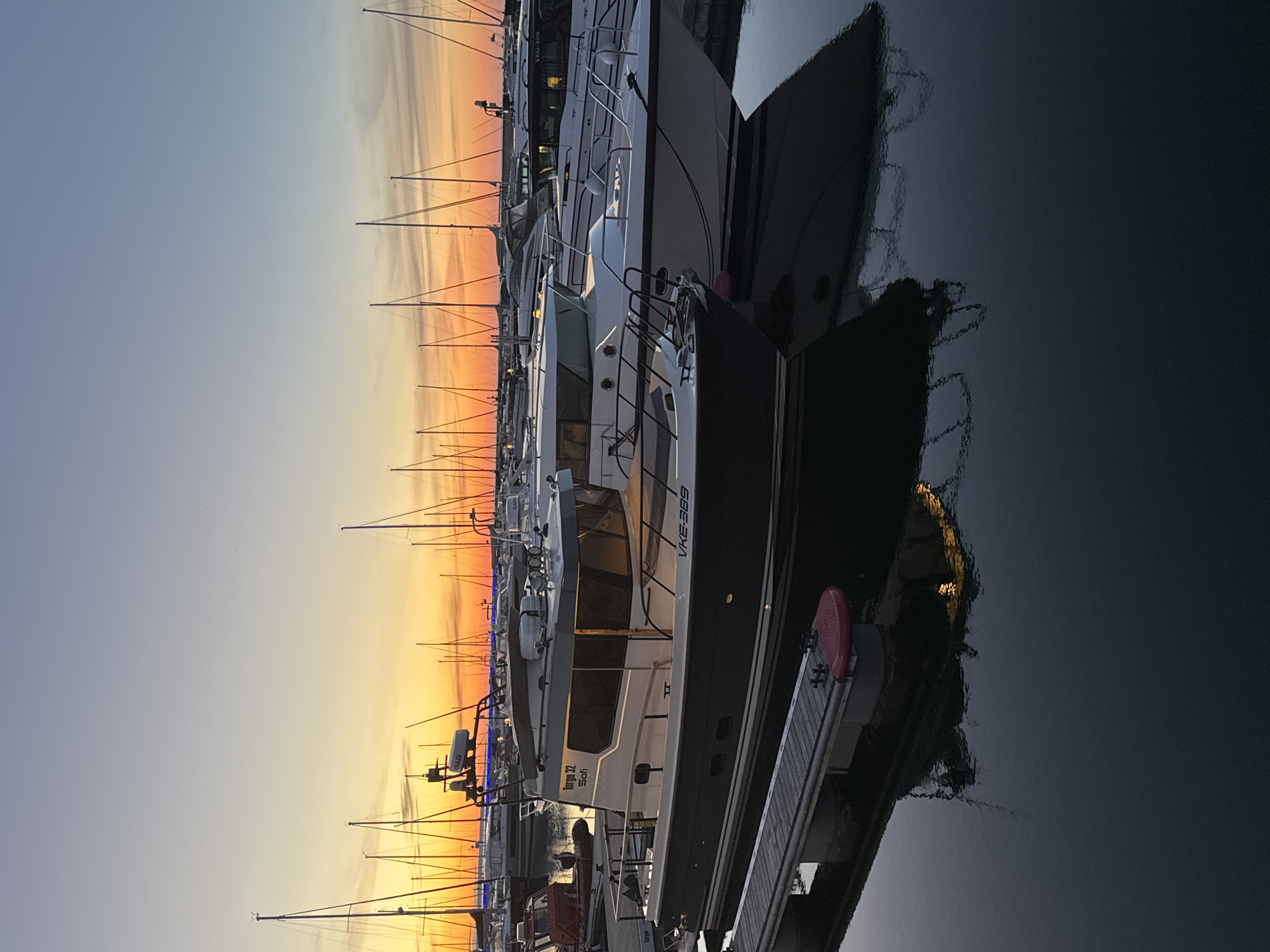 The image depicts a harbor scene at sunset in Tallinn, Estonia. The sky is a gradient of pale blue and vibrant orange, with thin clouds streaking across. Numerous sailboats with tall masts are docked in the background, silhouetted against the colorful sky. In the foreground, several motorboats are moored.
The most prominent boat in the foreground is a dark-hulled vessel with a white cabin. It has a railing around the deck and windows that reflect the sky. The name "Targa 32 Sofi" is visible on the cabin, and a registration number "VKE-389" is also present. A wooden pier with red fenders extends from the left side of the frame towards this boat.
Other motorboats are docked to the right and behind the main vessel. Some of these are white, with visible railings and cabin structures. The water in the harbor is dark and calm, reflecting the boats and the colorful sky. The overall impression is one of a peaceful evening in a well-maintained harbor. There are no people visible in the image.