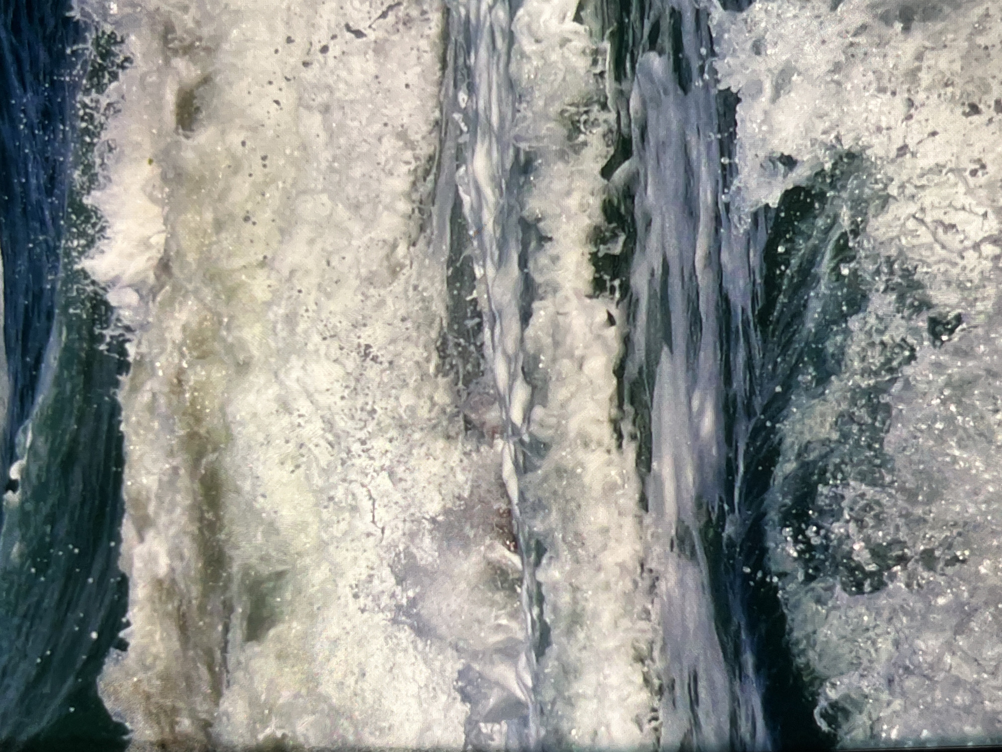 The image depicts a close-up view of turbulent ocean waves. The water is a deep blue-green, with white foam and spray churning as the waves crest and break. Sunlight glints off the water droplets, creating a shimmering effect. The texture of the water is rough and dynamic, suggesting a powerful and active sea. There are no visible people, structures, or landforms in the frame.