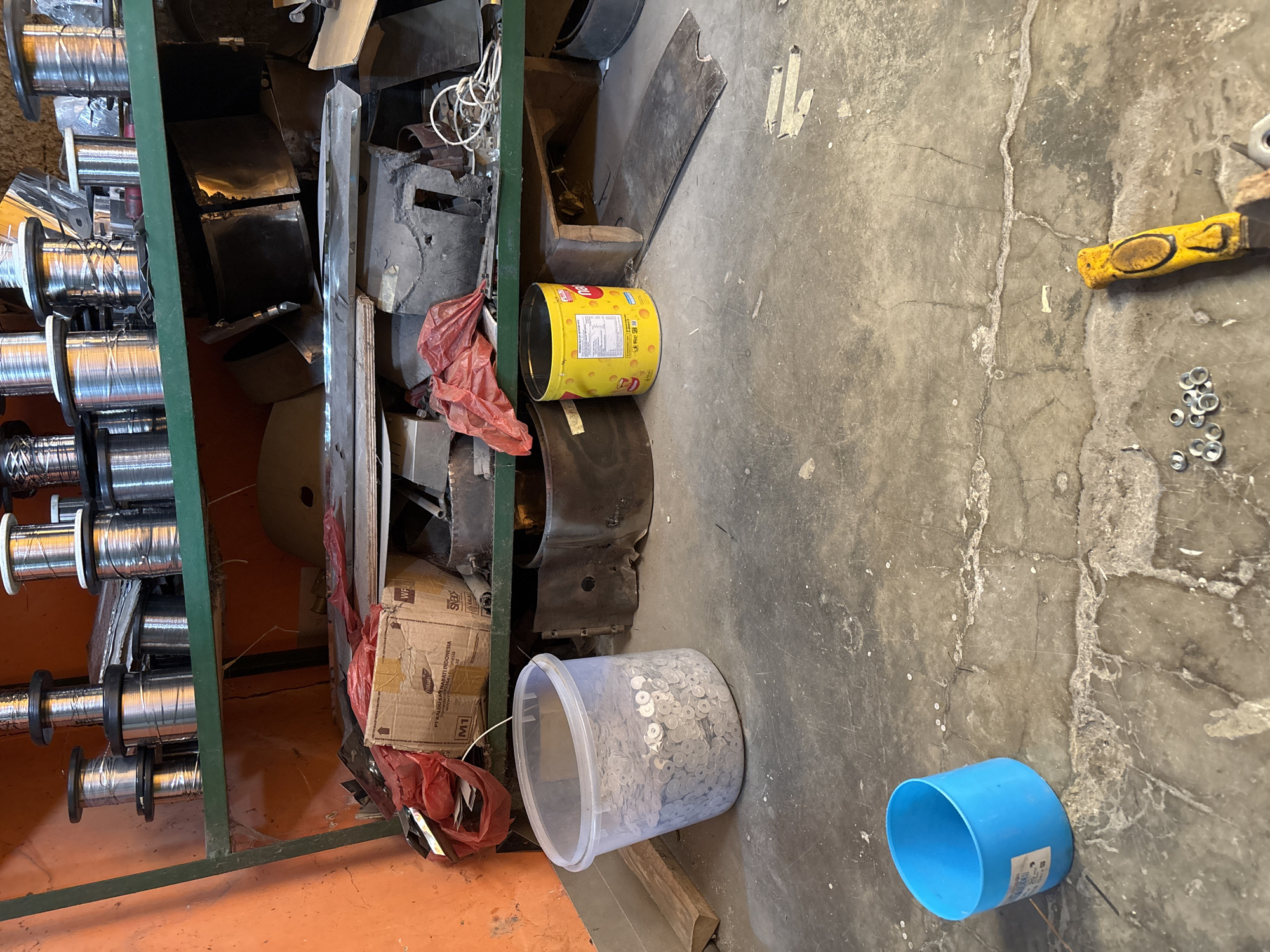 The image depicts a cluttered storage area, possibly a workshop or warehouse, located in Telagamurni, Indonesia. On a green metal shelf, spools of wire are neatly stacked. Below the shelf, various items are piled haphazardly, including a red plastic bag, a cardboard box with Indonesian text, and what appears to be metal sheeting. On the concrete floor, a clear plastic tub is filled with numerous small metal washers. To the right of the tub is a yellow cylindrical container with product labeling. Further to the right, there are metal cylindrical objects and loose metal sheets. In the foreground, a bright blue plastic cup is visible, and to the far right, a tool with a yellow handle lies on the floor near a small pile of washers. The overall environment appears to be a functional, albeit disorganized, space.