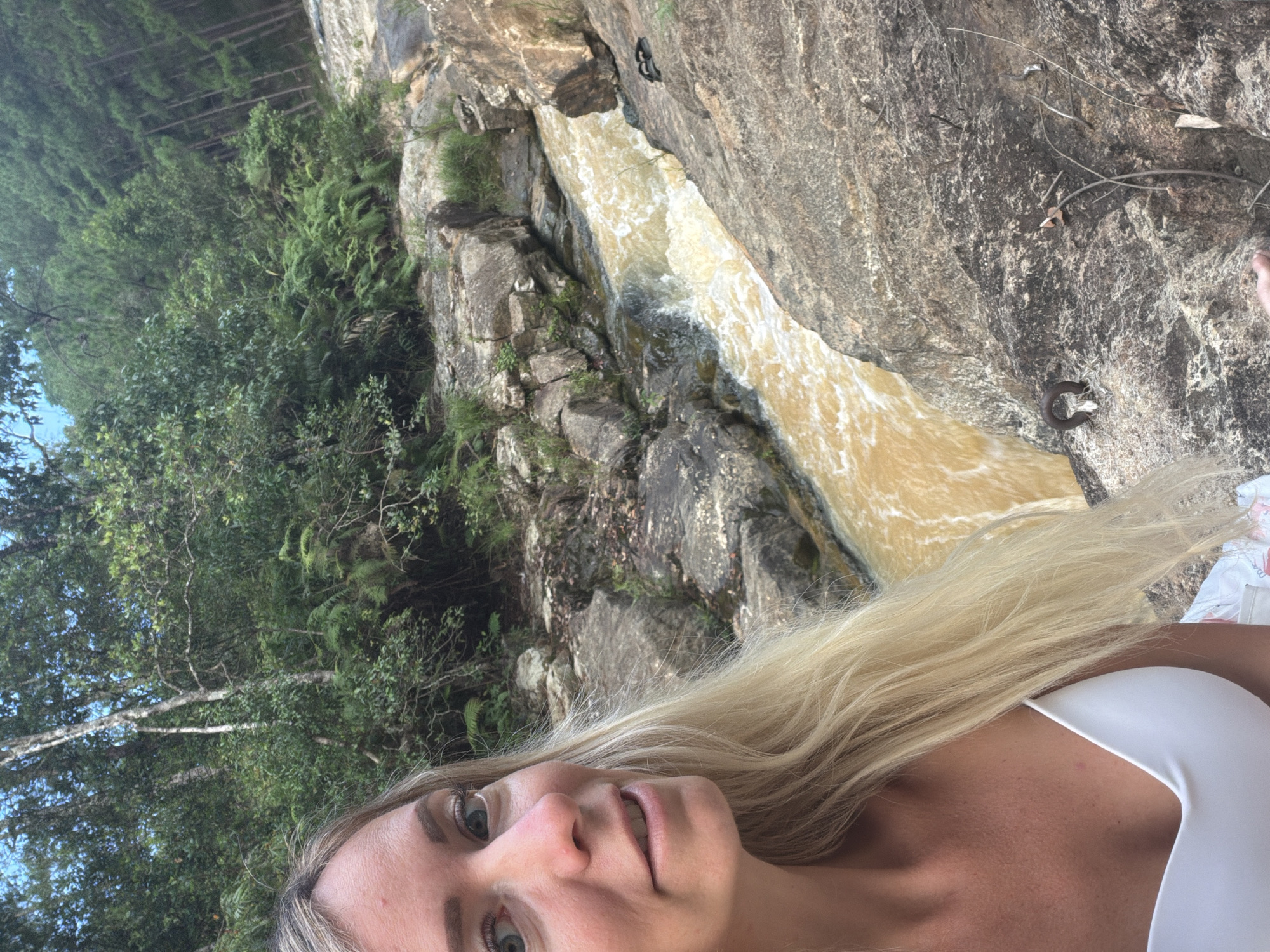 A woman with long blonde hair and blue eyes is taking a selfie near a rushing, muddy river. She is wearing a white bikini top. The river flows through a rocky canyon with lush green trees on either side. The trees on the right appear to be pine trees on a hillside. The river water is a murky brown, indicating it might be carrying sediment or is part of a larger, more turbid body of water. The rocks are rough and uneven, suggesting a natural, untamed landscape. The overall scene depicts an outdoor adventure setting, likely in a rural or natural area. There are no visible indicators that the location is in the city of Păng Tiêng 1, Vietnam.