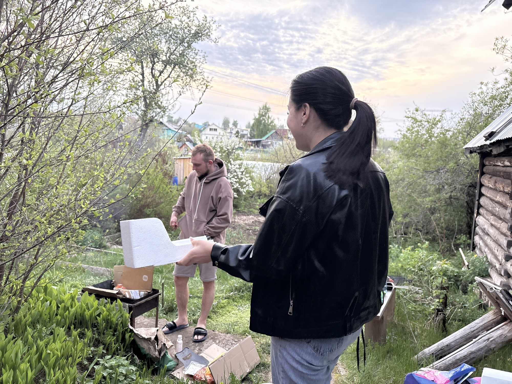 The image shows two individuals in an outdoor setting. One person, a woman with dark hair pulled back in a ponytail, is facing away from the camera, wearing a black leather jacket and light blue jeans. She is holding a piece of white polystyrene foam. The second person, a man, stands behind her near a charcoal grill. He is wearing a light brown hoodie and khaki shorts with sandals. He is looking down at the grill. Behind the grill is a cardboard box, suggesting they are preparing food.
The environment is a grassy area with various trees and bushes, indicating a backyard or garden setting. There is a log cabin structure on the right side of the image. In the background, there are houses and trees that suggest the location is a suburban or semi-rural area. Based on the visible landmarks and architectural style, the location is identified as Chaikovsky, Russia. The sky is overcast with a hint of sunset, indicating either dawn or dusk.