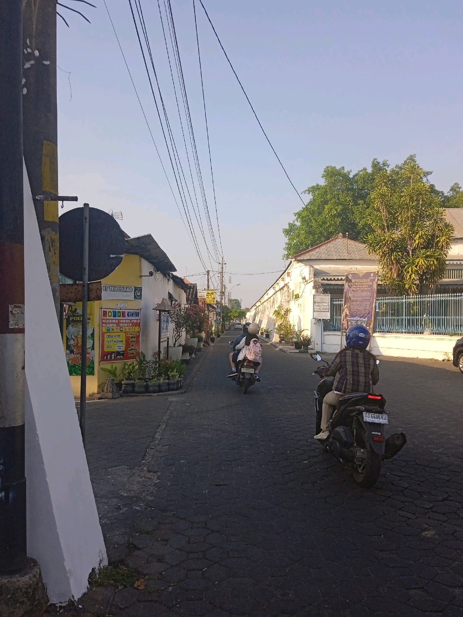 This is a street scene in Surakarta, Indonesia, captured in the late afternoon. The image looks down a paved street lined with low-rise buildings and trees. In the foreground, a black utility pole stands to the left, partially obscuring a white, angular structure. A few motorcycles are visible on the road. The first motorcycle, closer to the foreground, has two occupants, the passenger wearing a backpack. Further down the street, another motorcycle is ridden by a single person wearing a plaid shirt and a blue helmet.
The buildings on the left appear to be shops or small businesses, one of which has a colorful advertisement featuring a beverage called "ES TEH JUMBO" and another advertising a cell phone service and printing. The buildings on the right are whitewashed with tiled roofs. One prominent building on the right has a large blue and gold banner displaying text and images related to "REVITALISASI" and "KERATON" (palace). A blue metal fence runs along the front of this building. Overhead, a tangle of electrical wires stretches across the sky. The sky is clear and pale blue, indicating good weather. The road surface is made of dark, interlocking stones.