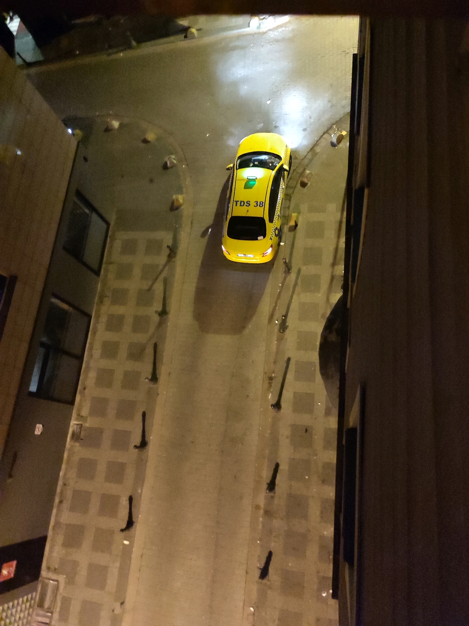 The image, taken at night from an elevated perspective, shows a yellow taxi with the license plate "TDS 38" parked on a street in Kadıköy, Turkey. The taxi is illuminated by its own headlights and overhead streetlights, casting shadows on the wet pavement. The street features a patterned tiled sidewalk and a narrow road. Adjacent to the street are building facades, one with visible windows. The scene appears to be an urban setting, possibly a quiet residential or commercial area, given the presence of the taxi and the architecture. There are no people visible in the image, and the taxi is stationary.