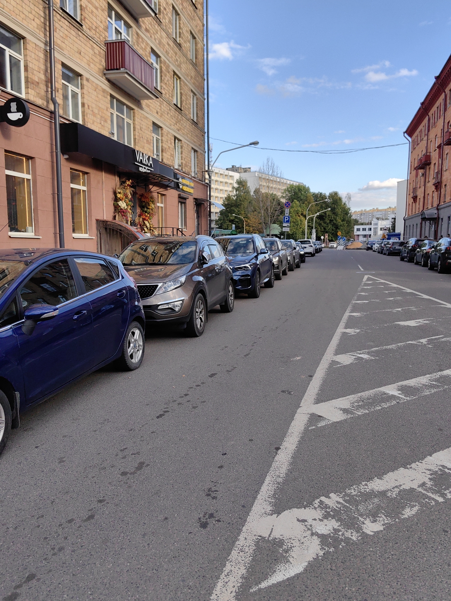 The image depicts a street scene in Minsk, Belarus, with cars parked along both sides of the road. On the left, a multi-story brick building with arched windows and a reddish-brown facade dominates the foreground. A black awning with the word "VARKA" in white letters is visible above the building's entrance. A blue car is parked in the immediate foreground on the left. On the right side of the street, a line of parked cars extends into the distance, with a red-brick building visible further down. The road is asphalt, with white lane markings and pedestrian crossing lines. The sky is blue with scattered clouds. Trees and other buildings are visible in the background, suggesting an urban environment. No people are clearly visible in the image.