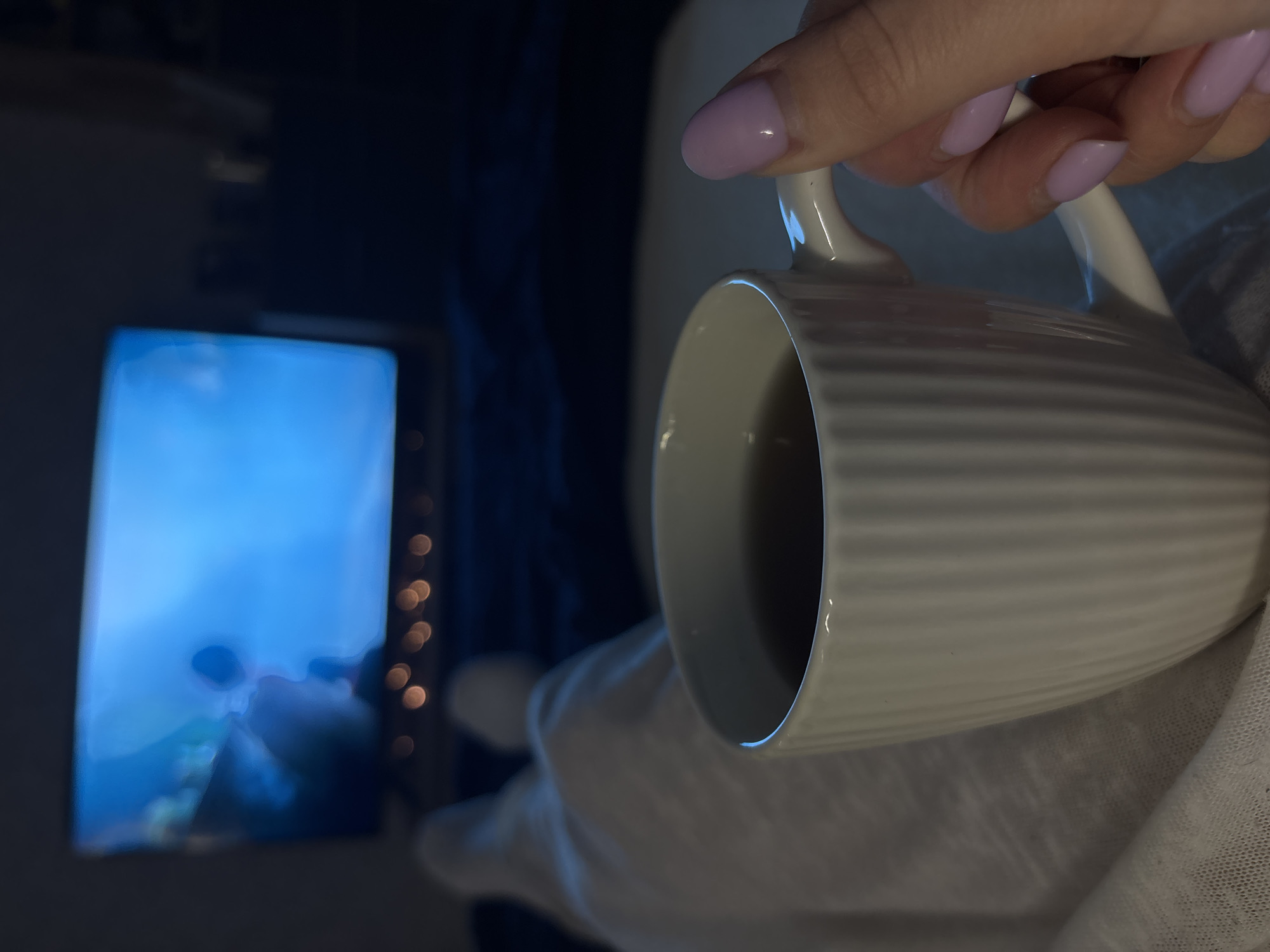 A close-up, angled shot features a hand holding a white, ribbed mug filled with a dark liquid, likely coffee or tea. The person has pale skin and painted nails in a light purple hue. The mug is positioned in the foreground, obscuring much of the background. In the blurred background, there is a television screen showing an underwater scene. To the right of the screen, faint lights are visible. White fabric covers the lower part of the image. The image does not show any elements that would make it possible to determine if it was taken in the city of Chaikovsky, Russia.