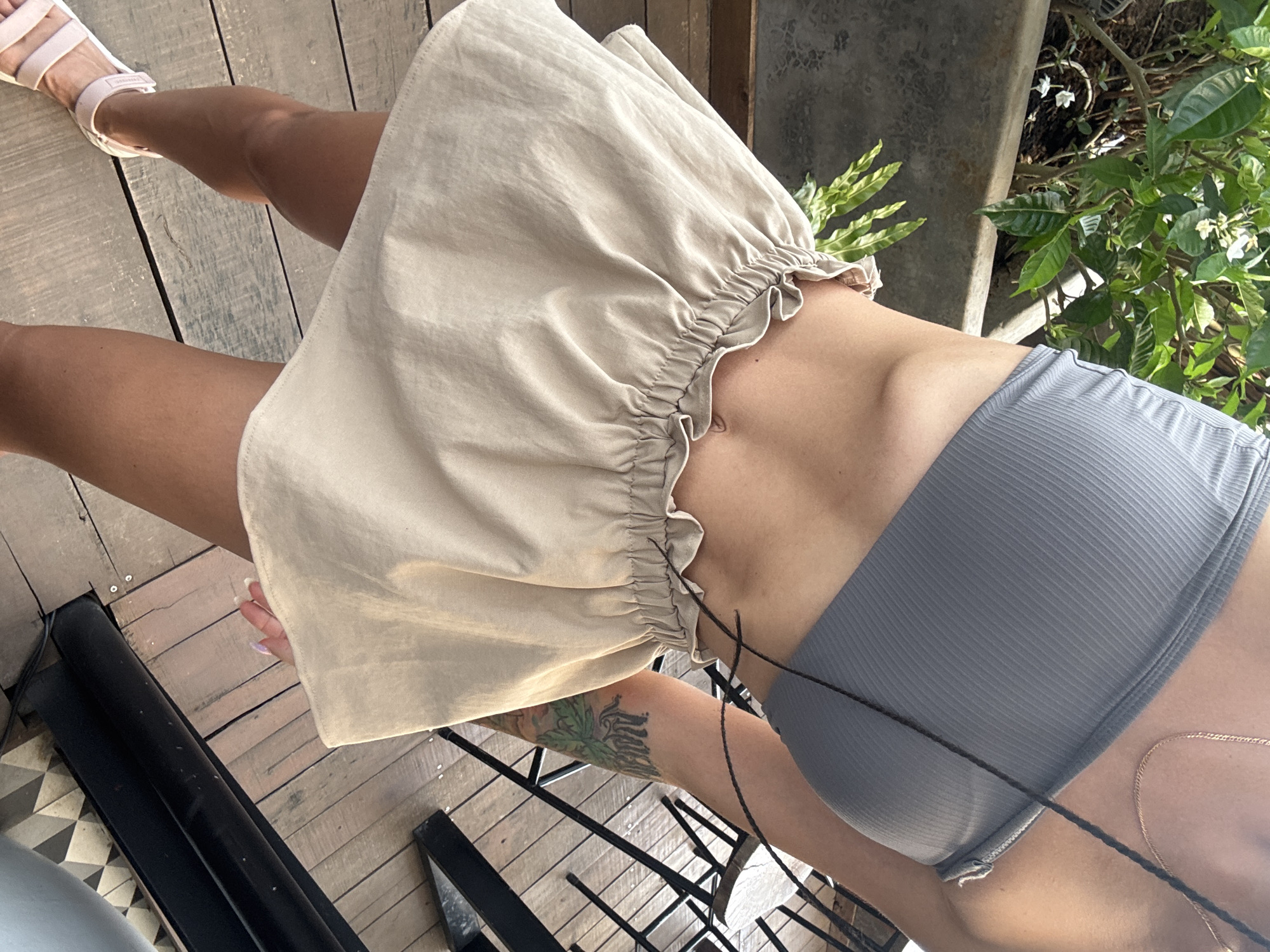A person, likely female, is partially visible in an outdoor setting, captured from a slightly elevated, downward-angled perspective. She is wearing a light beige or khaki-colored miniskirt with a ruffled, elasticated waistband and a grey ribbed bandeau top. Light pink flat sandals, with "ADIDAS" visible on a strap, are worn on one foot. A dark braided strap or string extends across the front of her torso, near the bandeau top. A colorful tattoo, featuring leafy patterns and dark script, is visible on her upper left leg. Her fingernails, partially seen near the skirt, are adorned with a light, possibly iridescent polish.
The person appears to be standing or posing casually. The setting includes a wooden deck floor composed of varied planks, along with some dark patterned tiles in the lower left corner. The background features lush green foliage, including bushes and plants with small white flowers, characteristic of a tropical or warm climate like Hà Giang 1 Ward, Vietnam. A concrete planter or structure and some metal railings are also present. The bright natural light indicates it is daytime, under sunny and clear weather conditions.