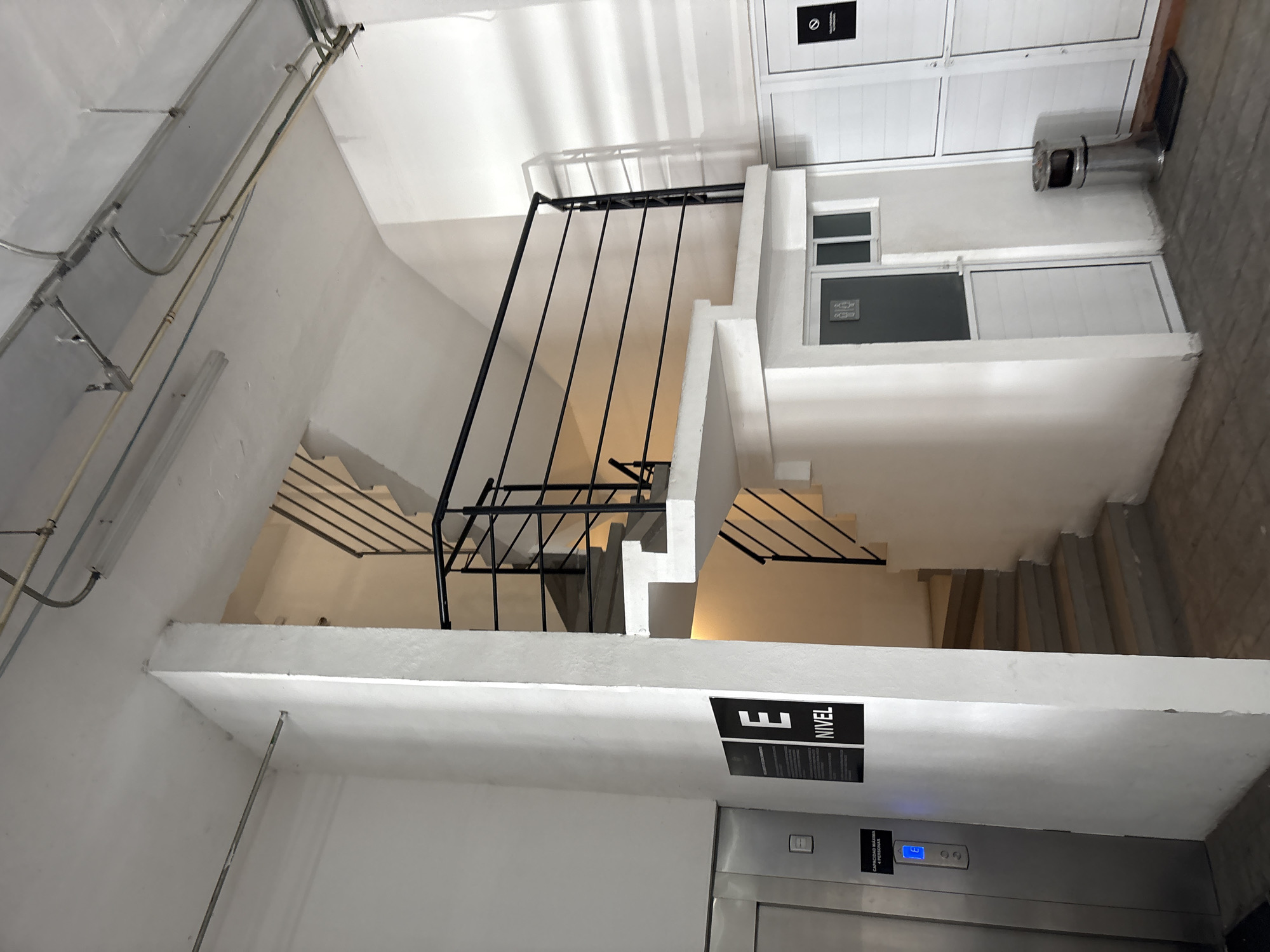 The image shows an interior view of a building in Puebla, Mexico. The scene features a stairwell with a black metal railing and white walls. Visible are electrical conduits running along the ceiling, an elevator with a button panel indicating the 3rd level, and signage in Spanish ("E NIVEL"). The stairs lead up to a landing with a window and door, along with a metal trash receptacle. The lighting appears to be a mix of natural light and artificial illumination.