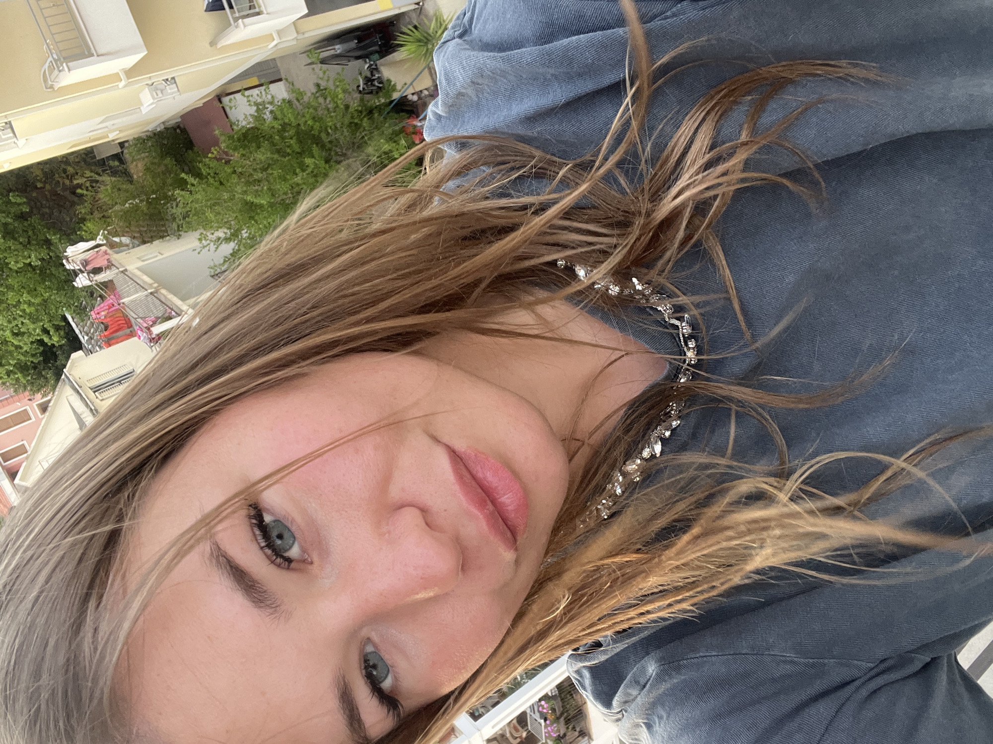 A close-up selfie of a young woman with blonde hair, blue eyes, and fair skin. She is wearing a denim shirt and a silver necklace. The background shows an outdoor setting with a building, greenery, and what appears to be a balcony or terrace railing. The location is possibly Budva, Montenegro based on the architecture and vegetation in the blurred background. The photo is taken at an angle, with the camera positioned slightly above and to the side of the subject.